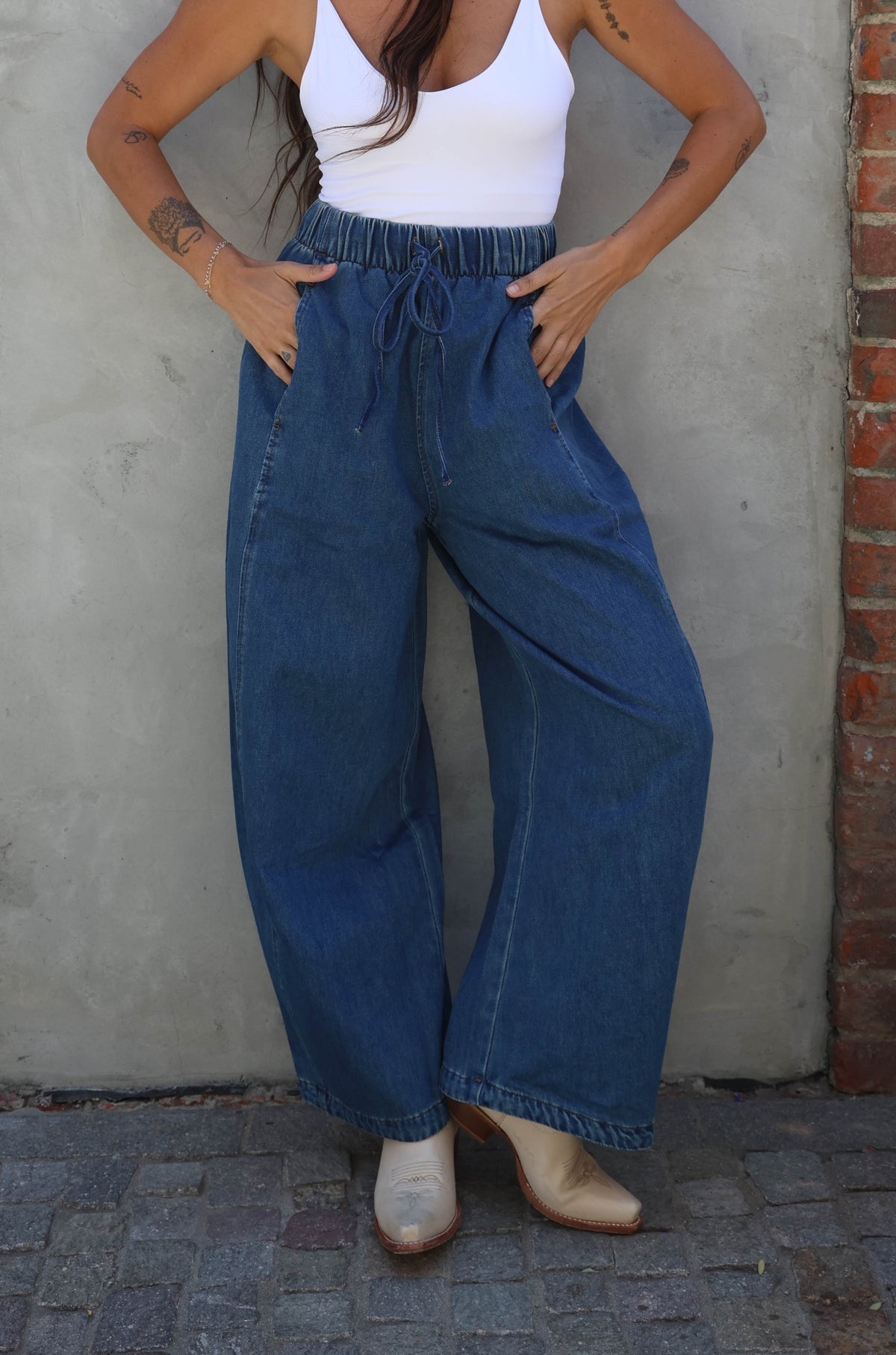 Woman wearing a white top and blue jeans standing against a brick wall.