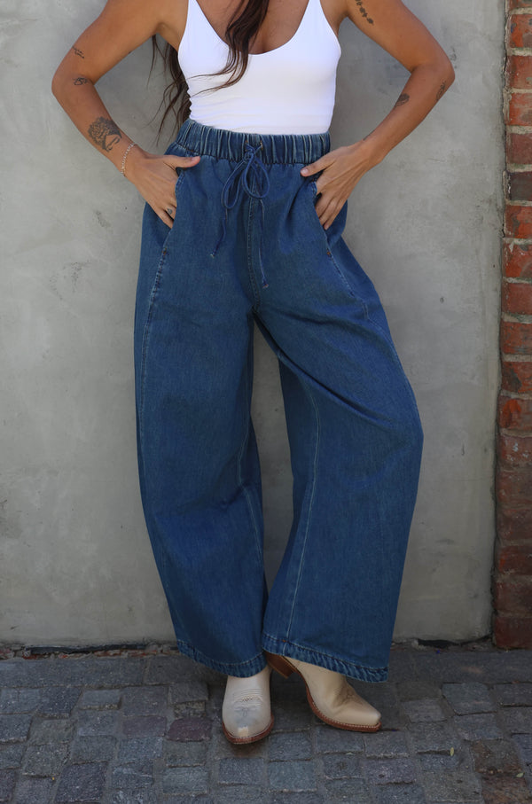 Woman wearing a white top and blue jeans standing against a brick wall.