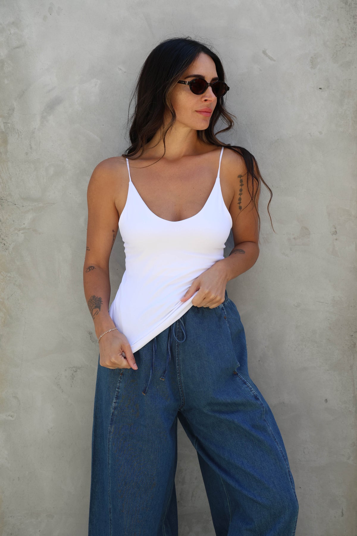 Woman wearing a white tank top and blue jeans against a gray wall