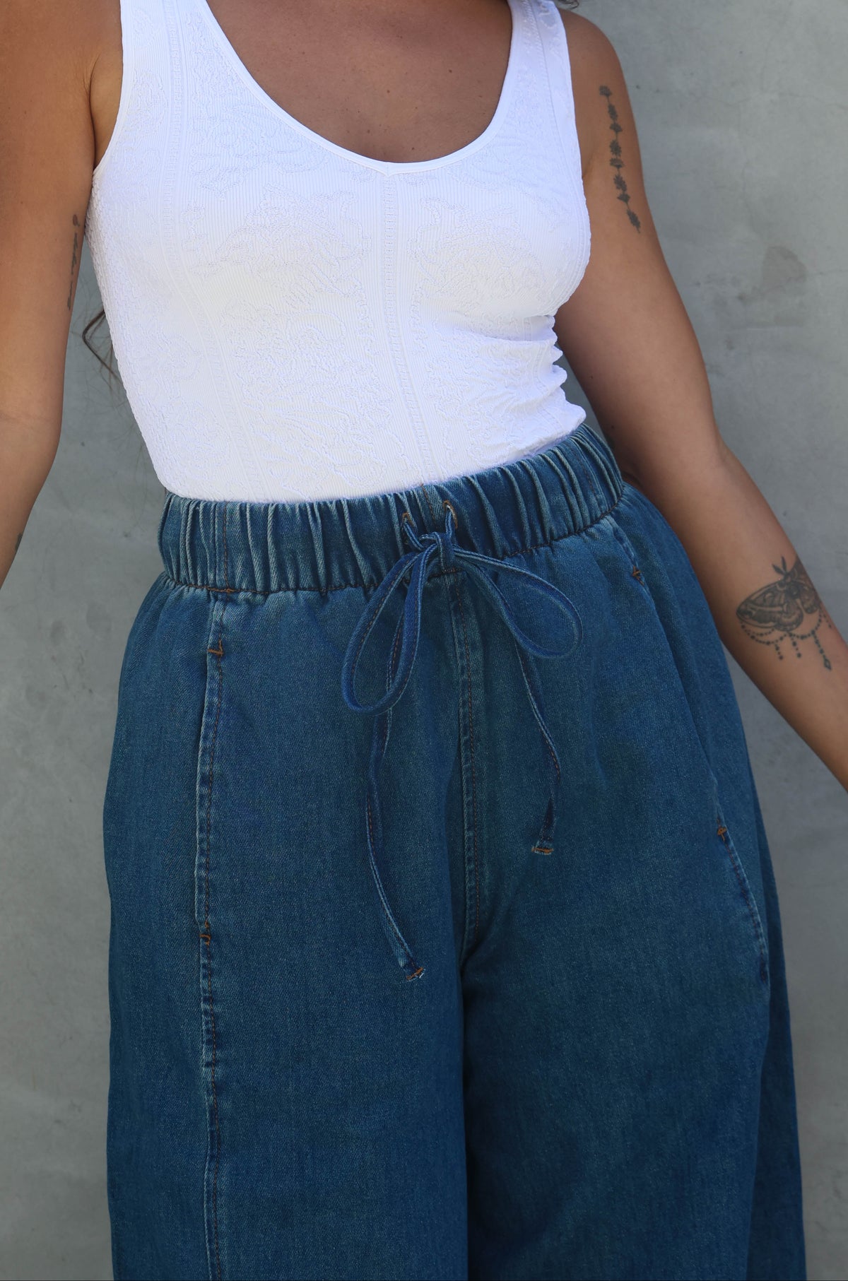 Woman wearing a white tank top and blue jeans against a gray wall.