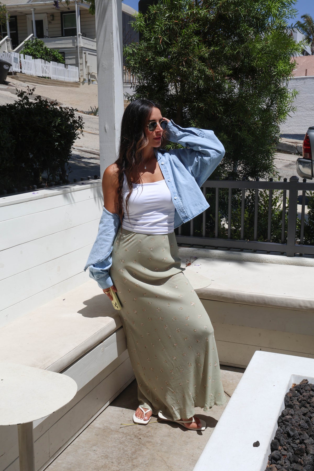 A woman posing outdoors on a patio wearing a white tank top, a light blue cropped denim shirt, and a long olive green skirt with small white polka dots.