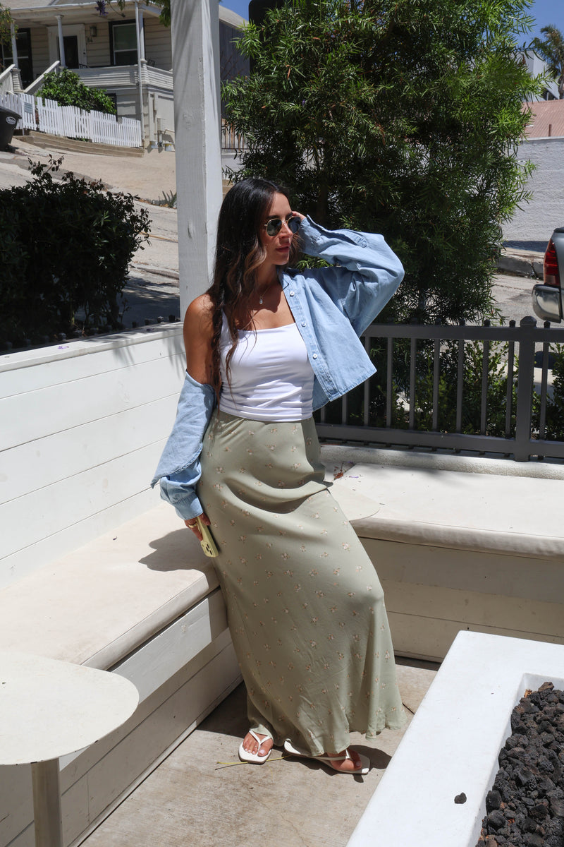 A woman posing outdoors on a patio wearing a white tank top, a light blue cropped denim shirt, and a long olive green skirt with small white polka dots.