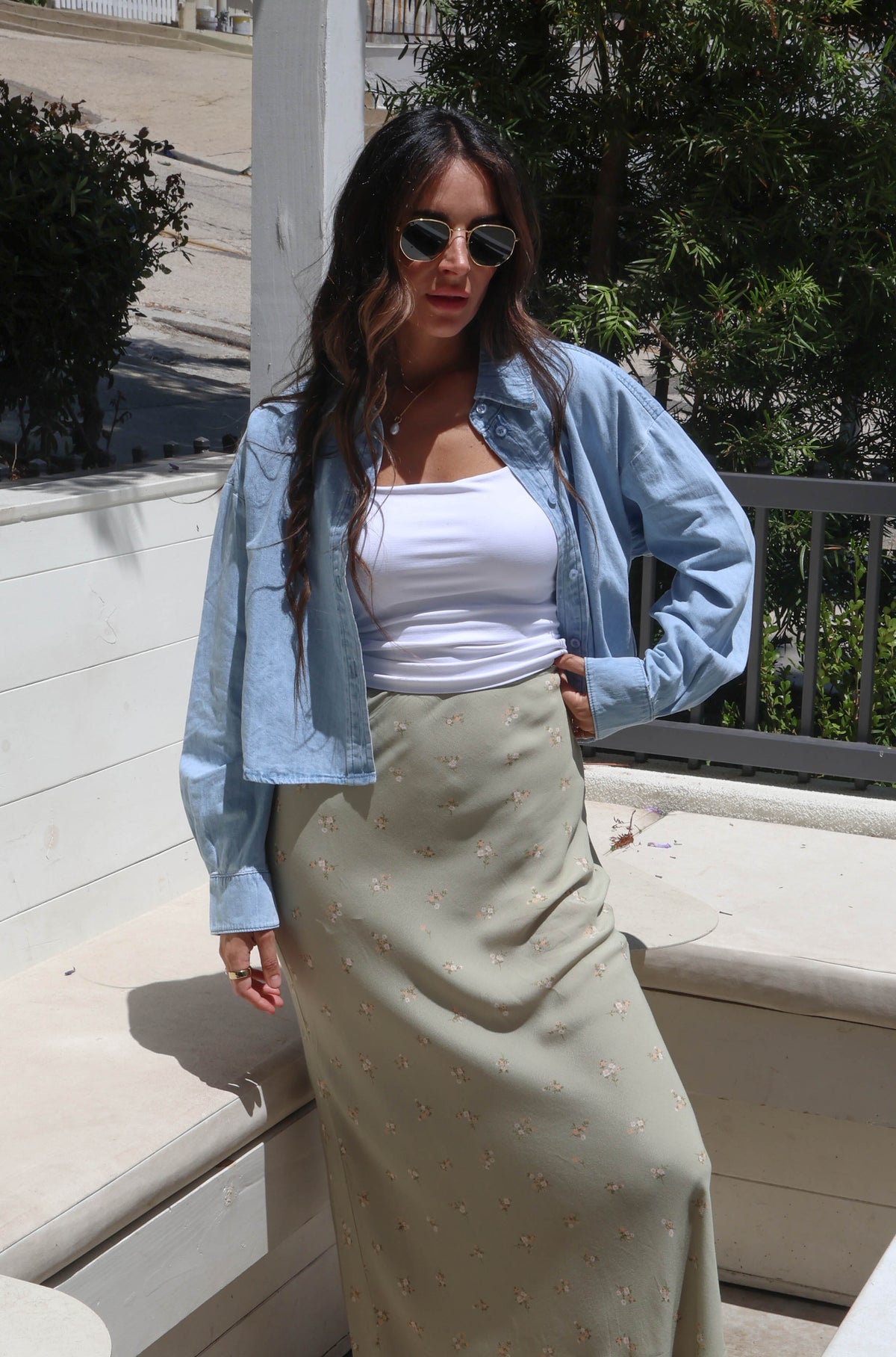 A woman stands outdoors on a patio wearing a light blue cropped denim jacket over a white tank top and a long beige maxi skirt. She is wearing sunglasses and white slide sandals, with a white bench, trees, and a house visible in the background.