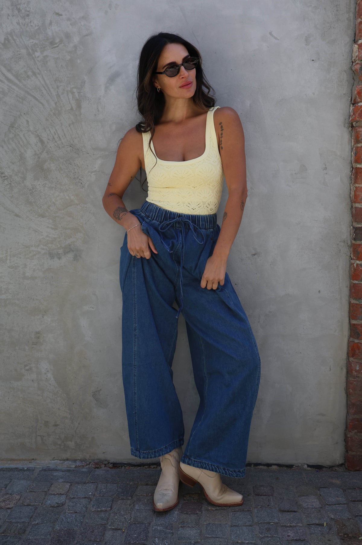 Woman wearing a yellow top and blue jeans standing against a textured wall.