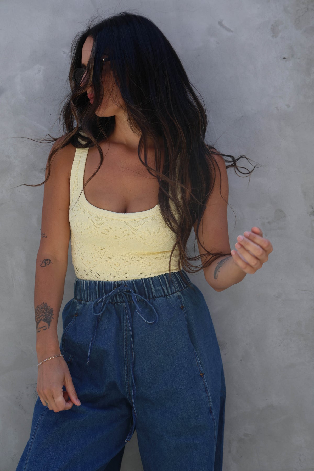 Woman wearing a yellow tank top and blue jeans against a gray wall