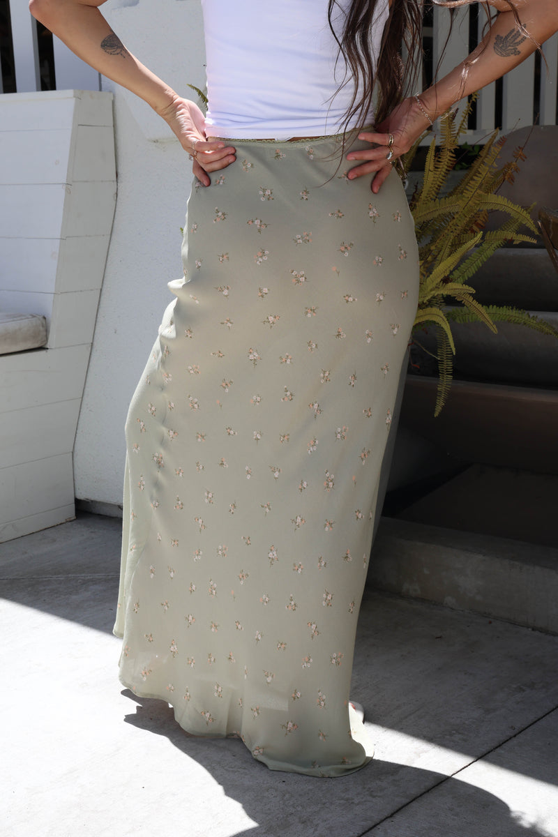A woman wearing a long, flowing sage green skirt with small embroidered floral details, paired with a white top. She is standing outdoors on a concrete patio with white steps and a plant in the background.
