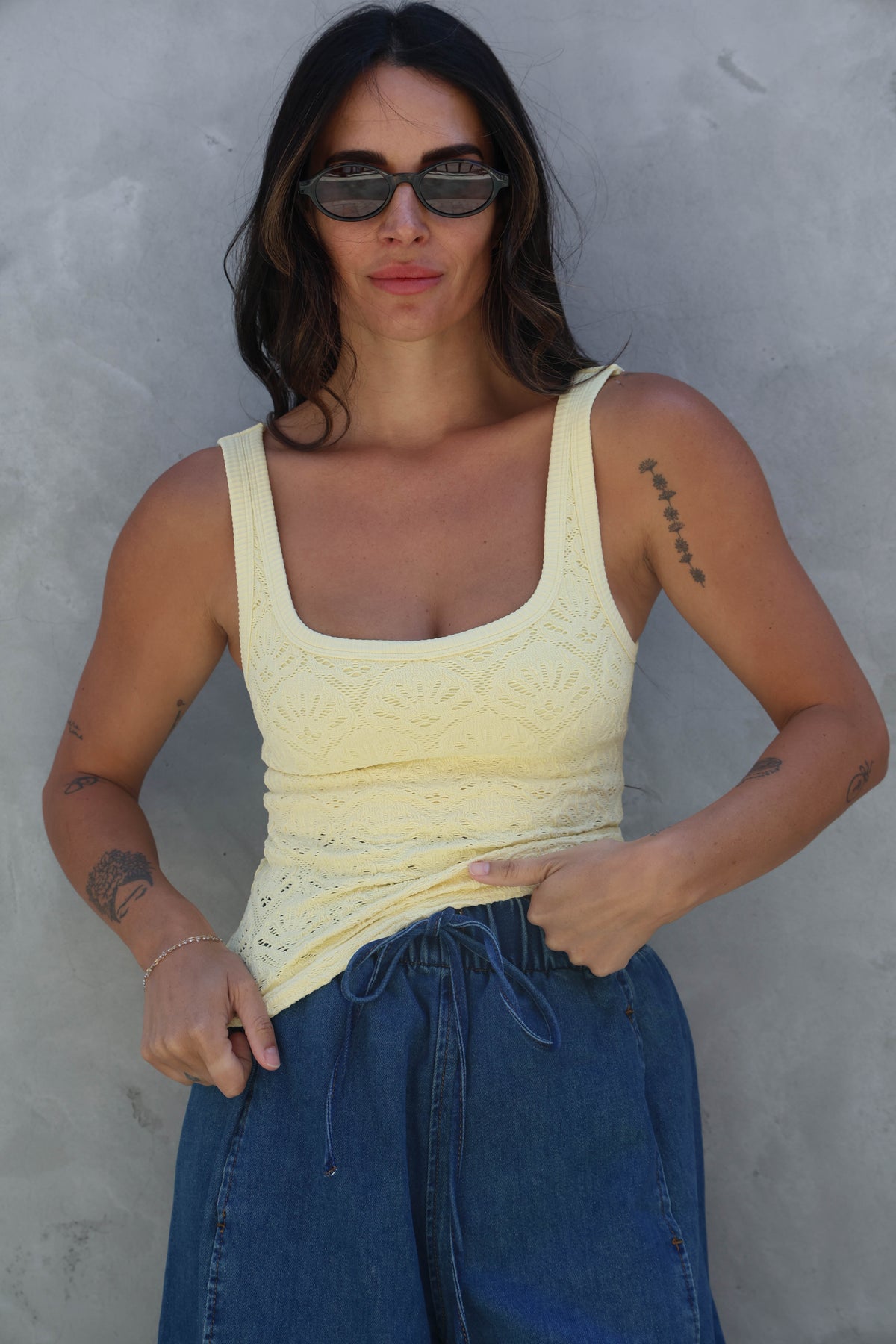 Woman wearing a yellow tank top and blue jeans against a gray wall.