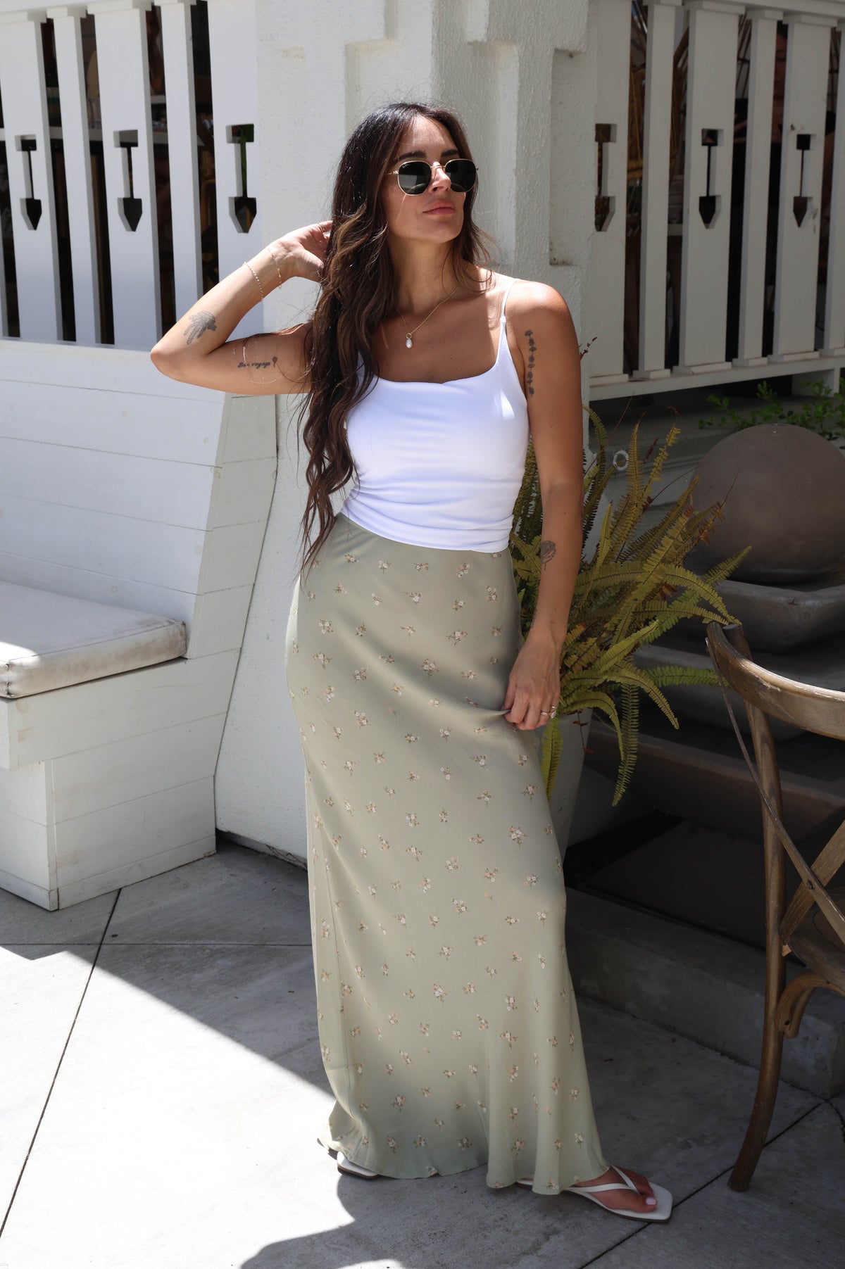 A woman wearing a white ribbed tank top and a long, light sage green skirt with small gold floral embroidery, posing on an outdoor patio.