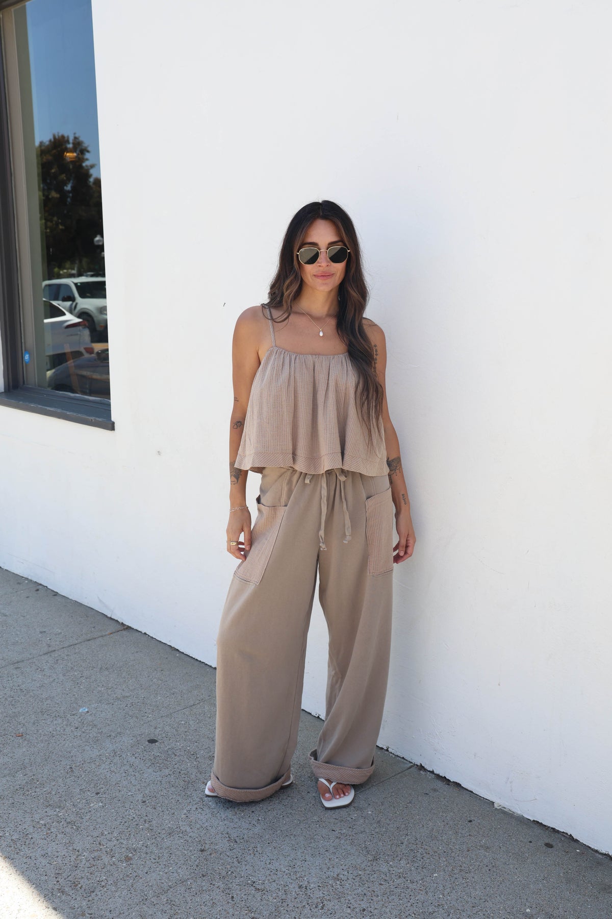 A woman wearing a beige two-piece outfit consisting of a ruffled strapless top and matching wide-leg pants, standing against a white wall outdoors.