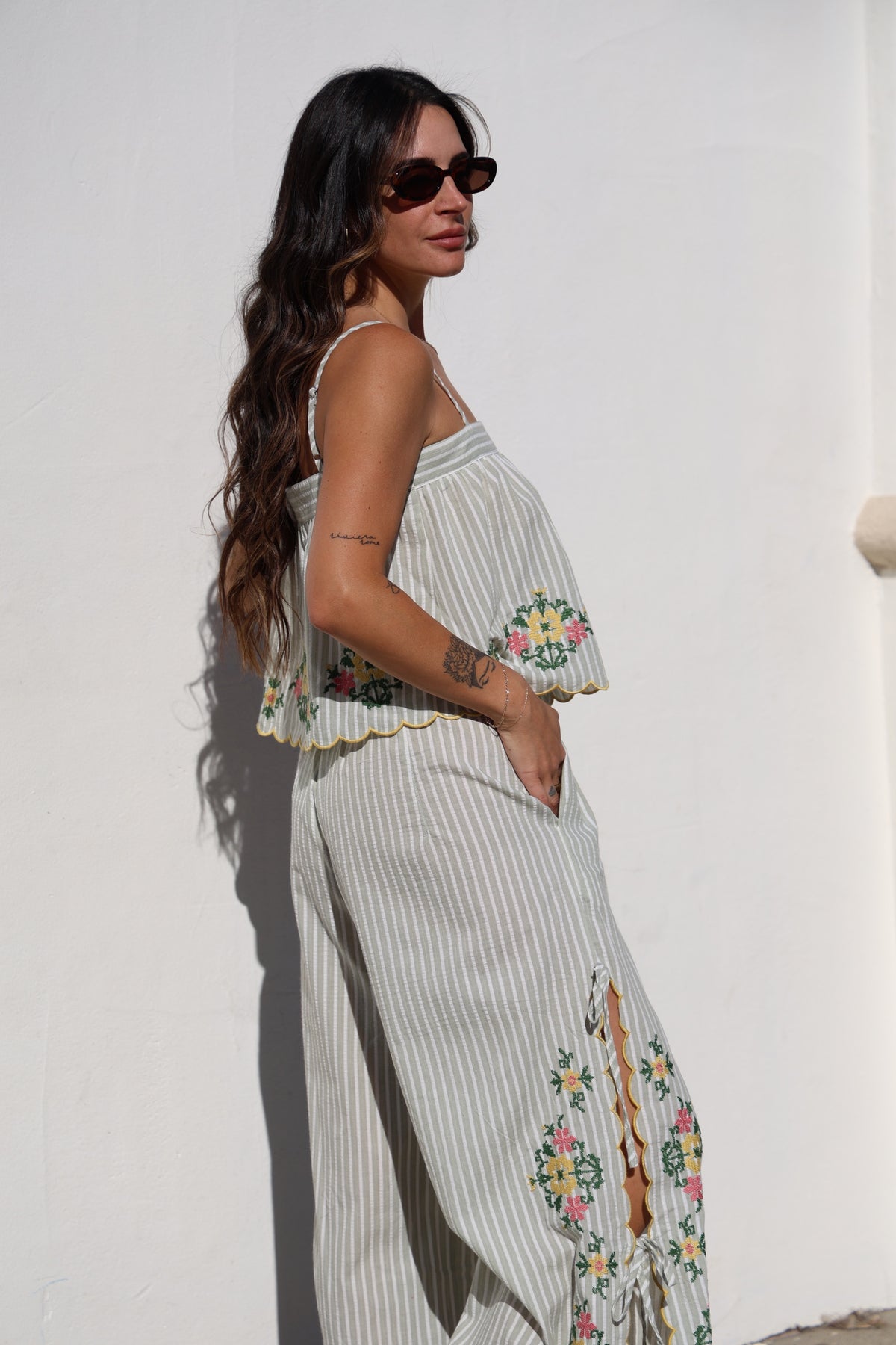 Woman wearing a long, light-colored dress with floral embroidery against a white wall.