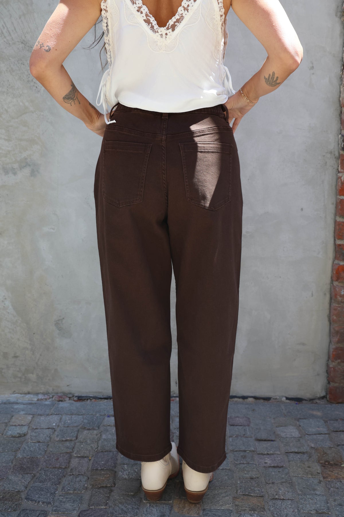 Person wearing a white top and brown pants with a visible tear, standing against a gray wall.