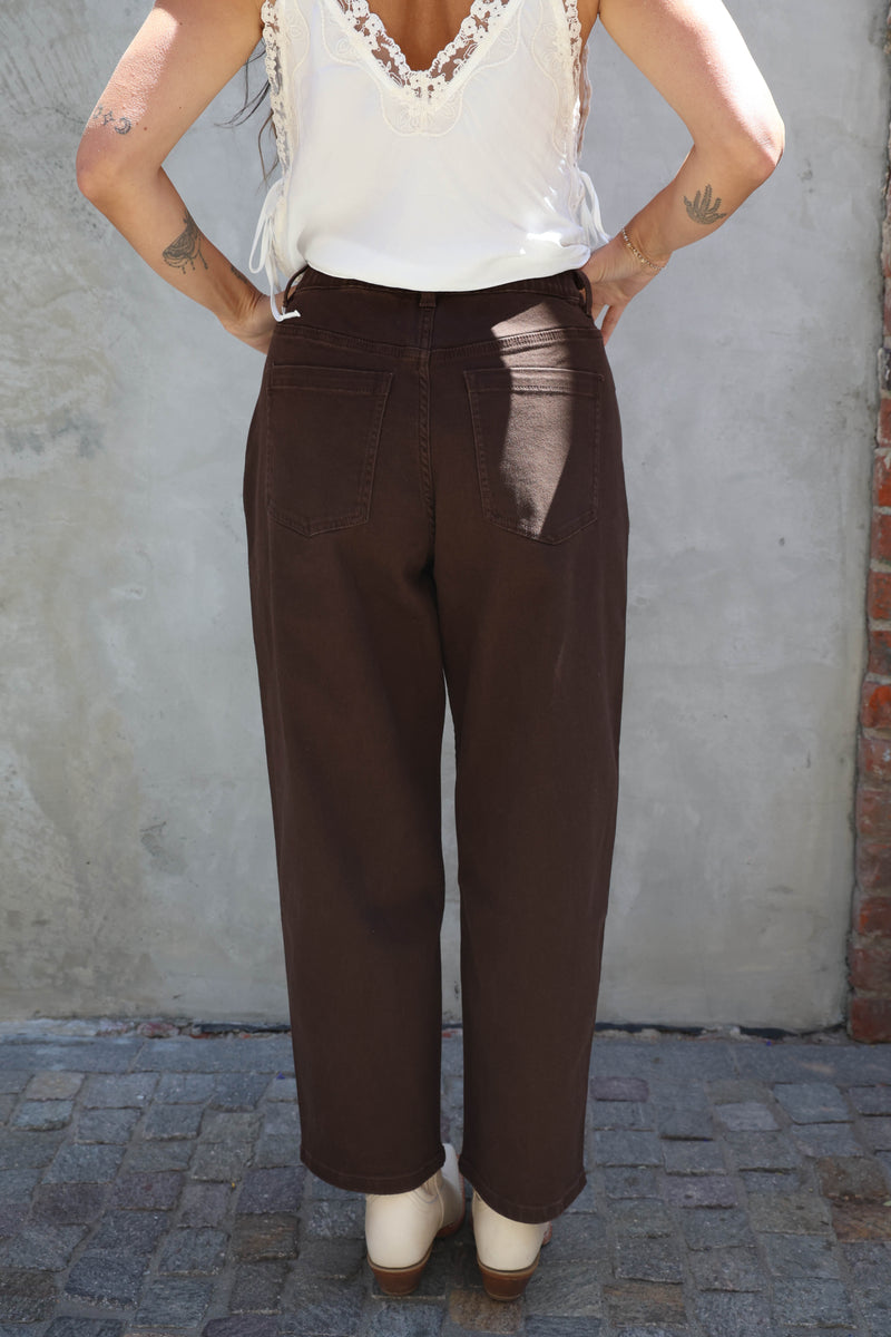 Person wearing a white top and brown pants with a visible tear, standing against a gray wall.
