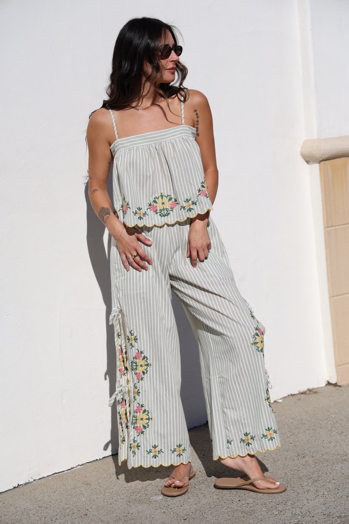 Woman wearing a light-colored embroidered jumpsuit against a white wall.