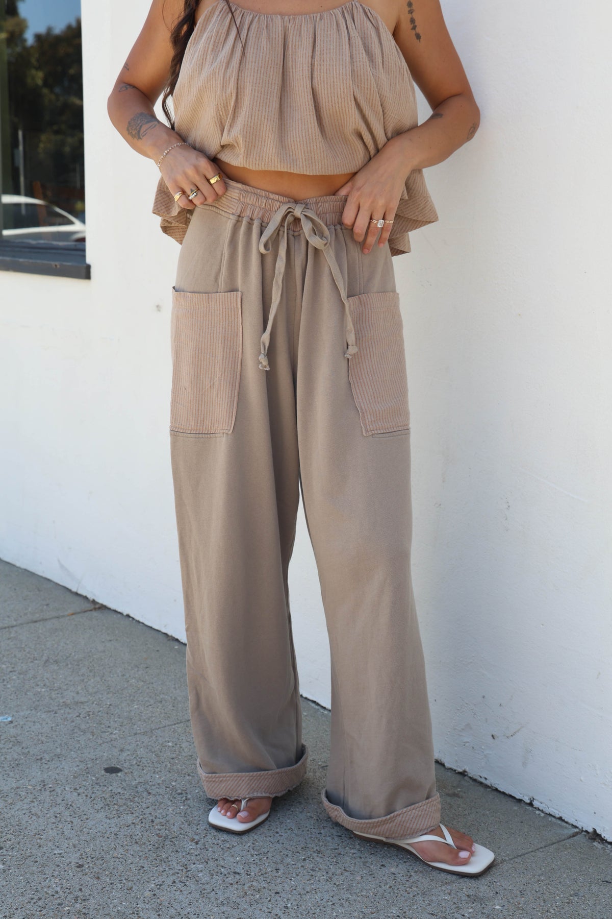A person wearing a beige two-piece outfit consisting of a sleeveless ruffled crop top and matching wide-leg drawstring pants with large patch pockets, standing outdoors against a white wall.