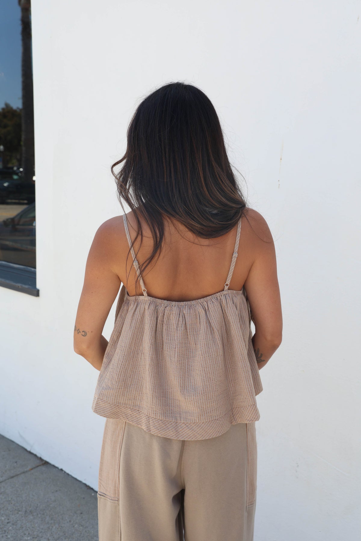A woman viewed from behind wearing a beige textured ruffled camisole top with thin straps and matching beige wide-leg pants, standing against a white wall.