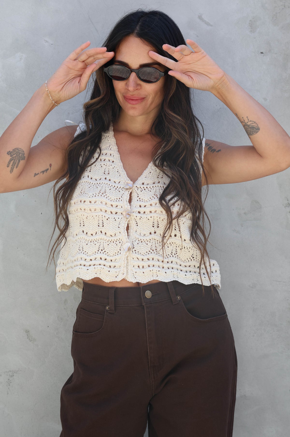 Woman wearing a white lace top and brown pants against a gray wall.