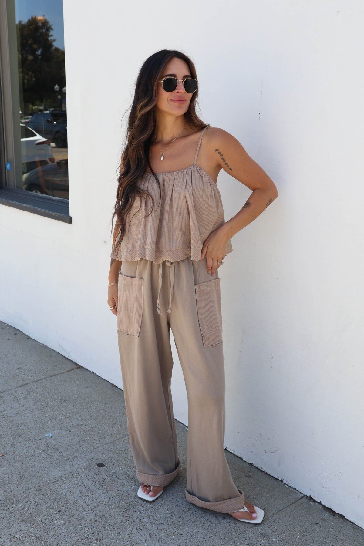 A woman wearing a matching beige two-piece set consisting of a sleeveless ruffled camisole top and wide-leg cargo pants, standing outdoors against a white wall.