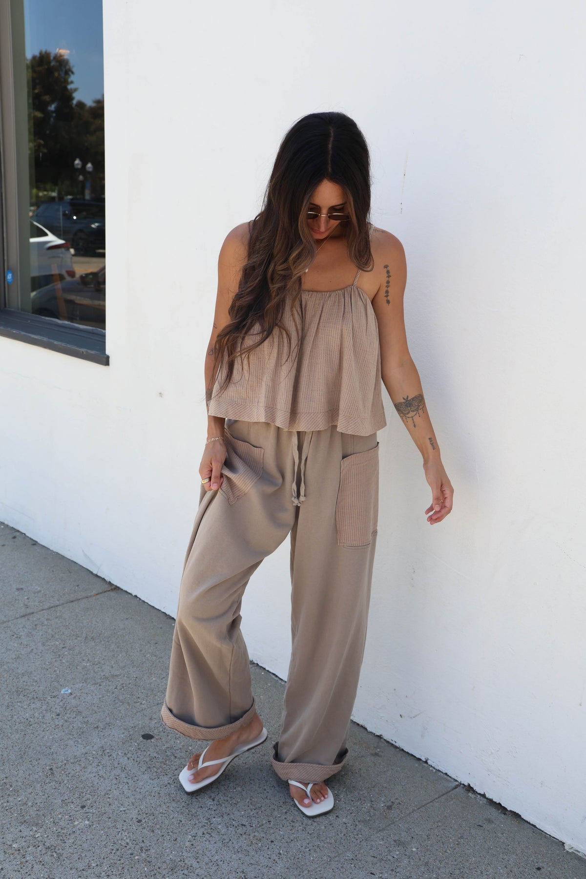 A woman wearing a beige two-piece lounge set consisting of a sleeveless ruffled top and matching wide-leg drawstring pants, standing outdoors against a white wall.