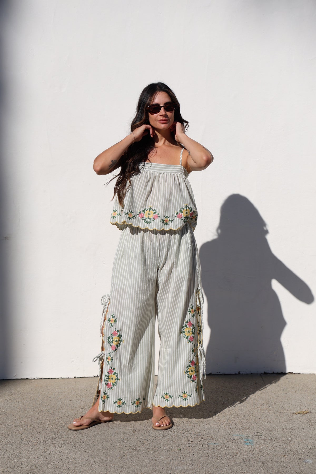 Woman wearing a light-colored outfit with floral embroidery against a white wall.