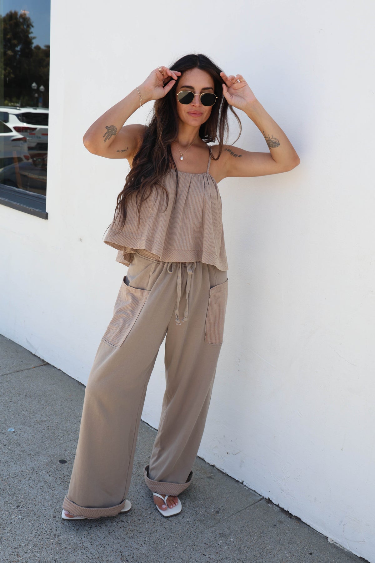 A woman wearing a matching beige two-piece lounge set consisting of a sleeveless ruffled top and wide-leg drawstring pants, standing outdoors against a white wall.