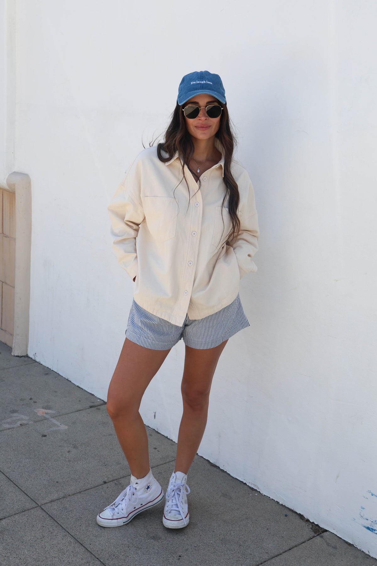 A woman stands against a white wall wearing an oversized cream-colored button-up shirt, light gray denim shorts, white sneakers, a blue baseball cap, and sunglasses.