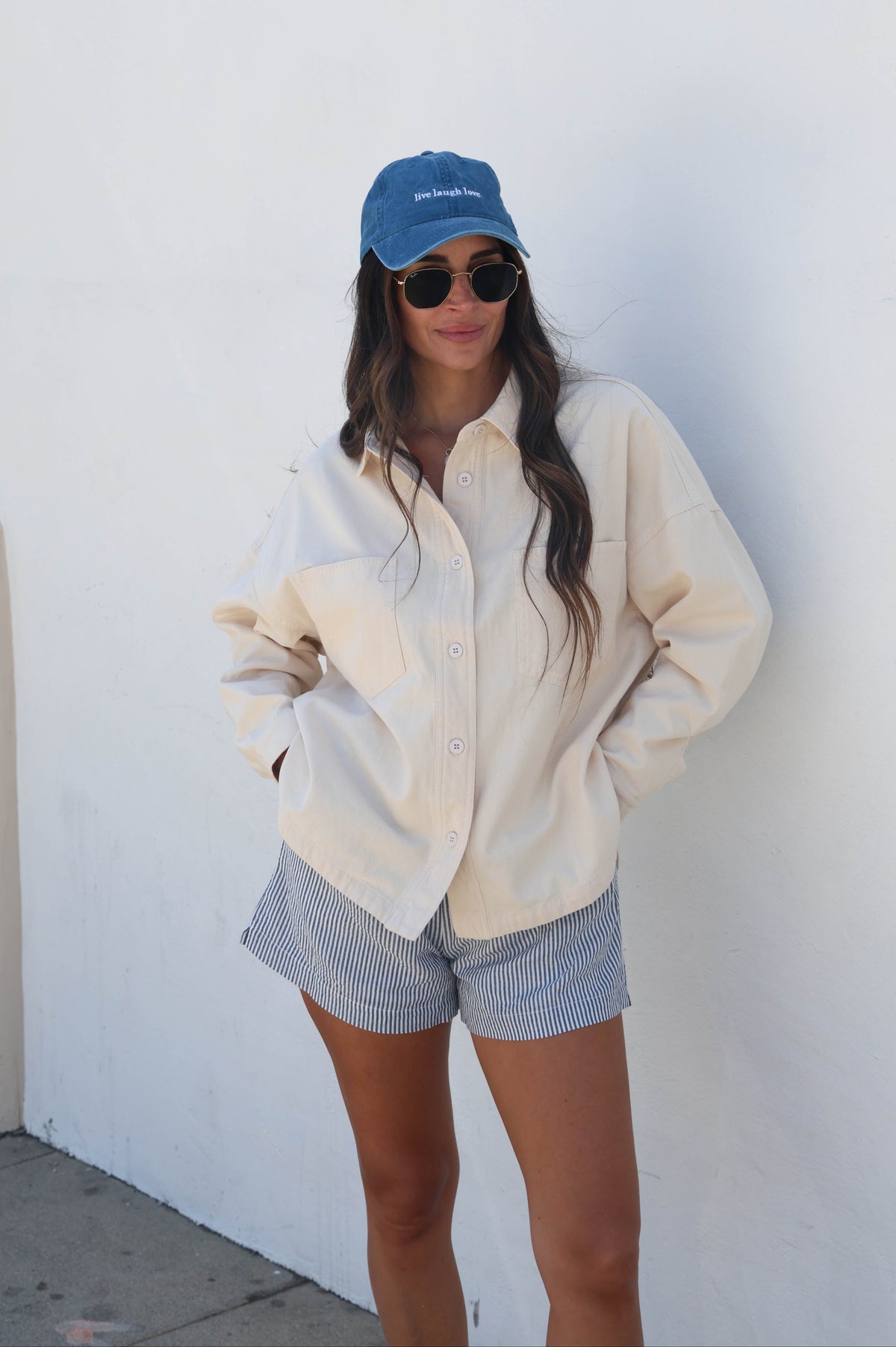 A woman stands against a white wall wearing an oversized cream button-up shirt, blue-and-white striped shorts, a blue baseball cap, and round sunglasses.