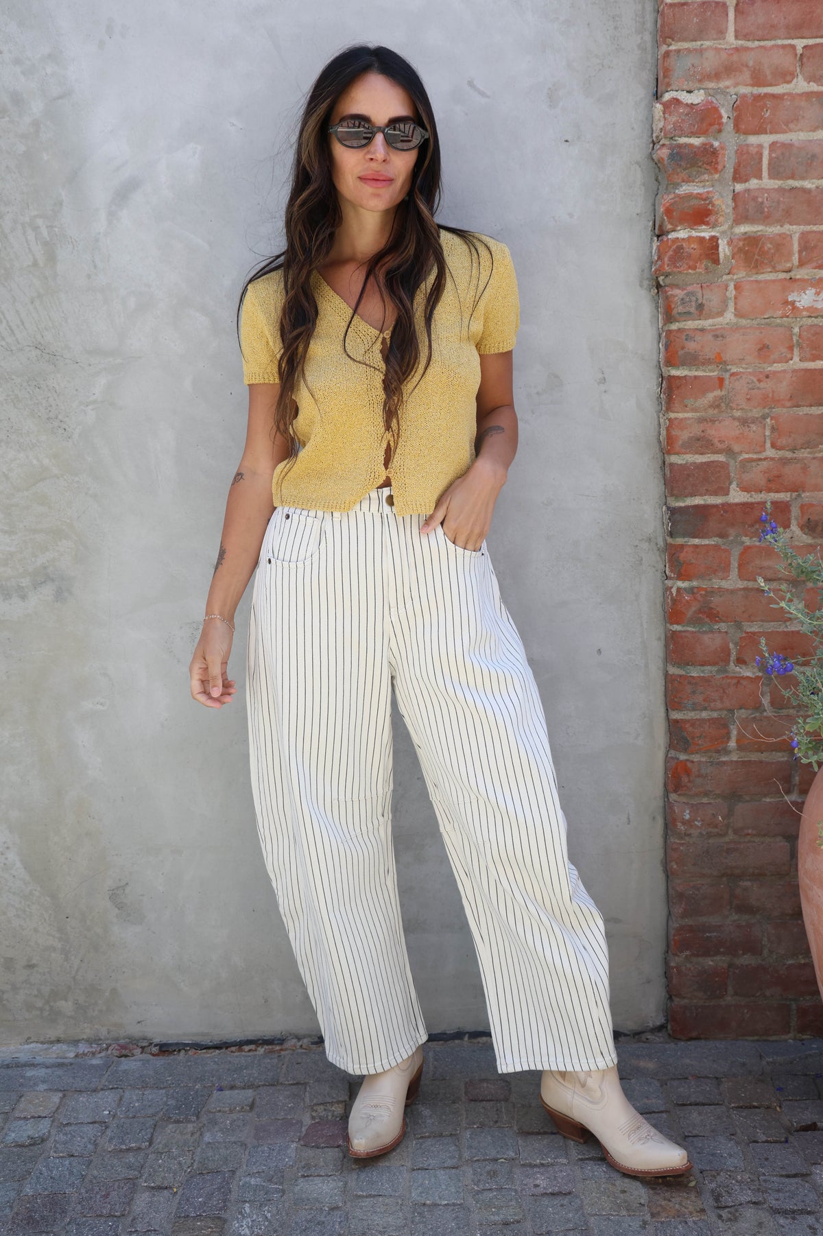 Woman in yellow top and white striped pants standing against a textured wall with a brick pattern.
