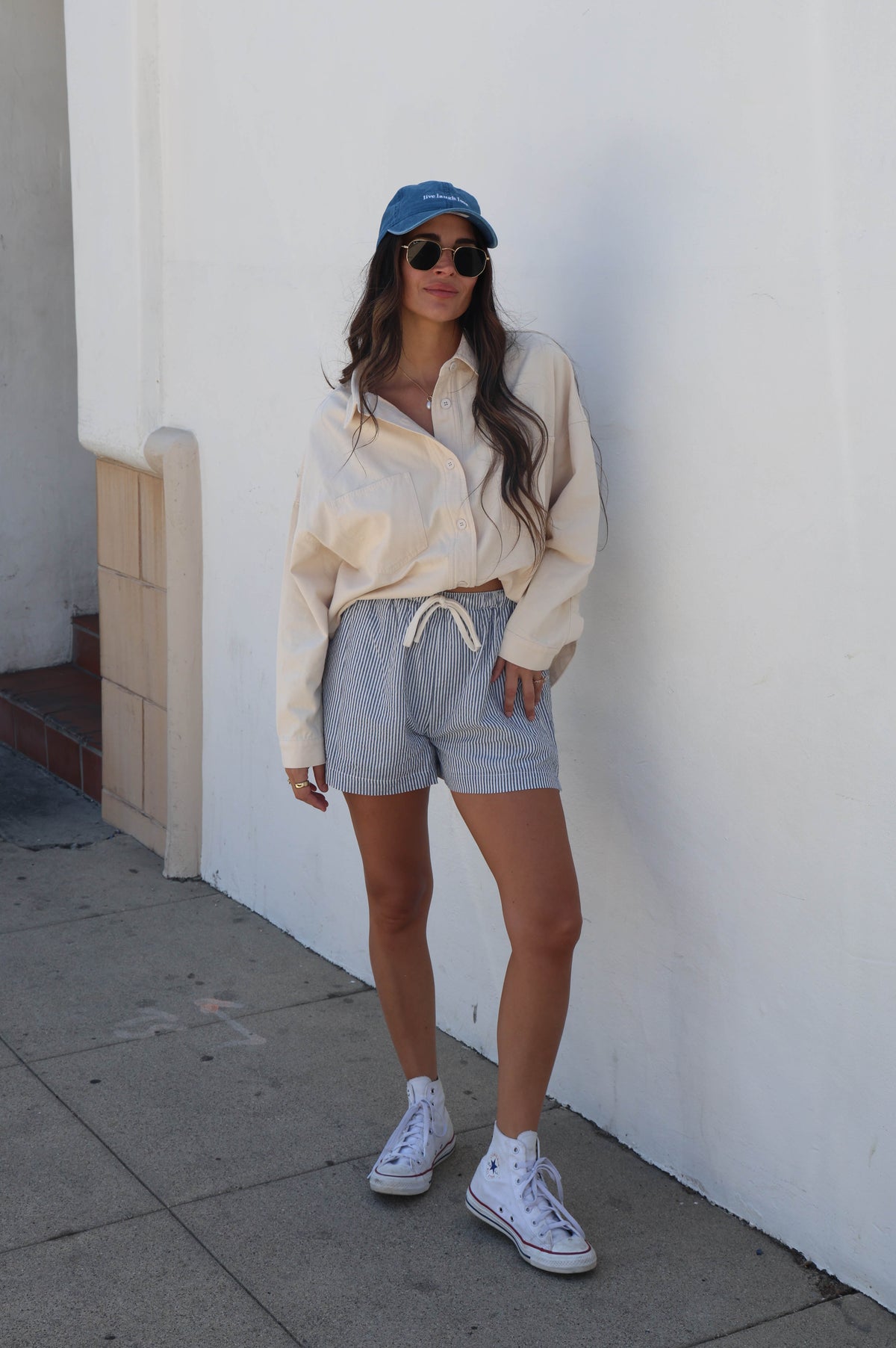 A woman stands against a white stucco wall wearing a cream button-up shirt, light blue drawstring shorts, white sneakers, white socks, and a blue baseball cap.