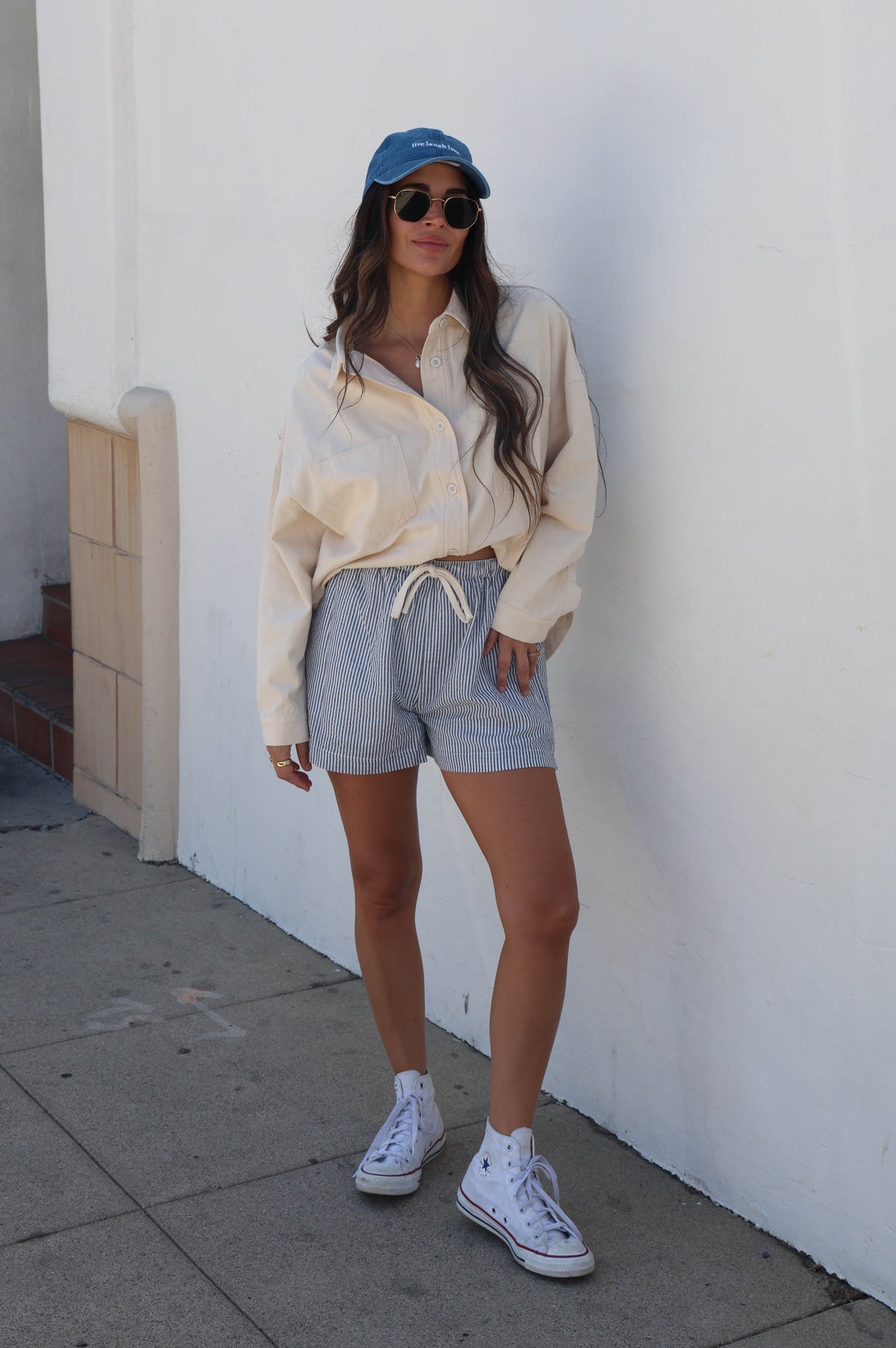 A woman stands against a white stucco wall wearing a cream button-up shirt, light blue drawstring shorts, white sneakers, white socks, and a blue baseball cap.