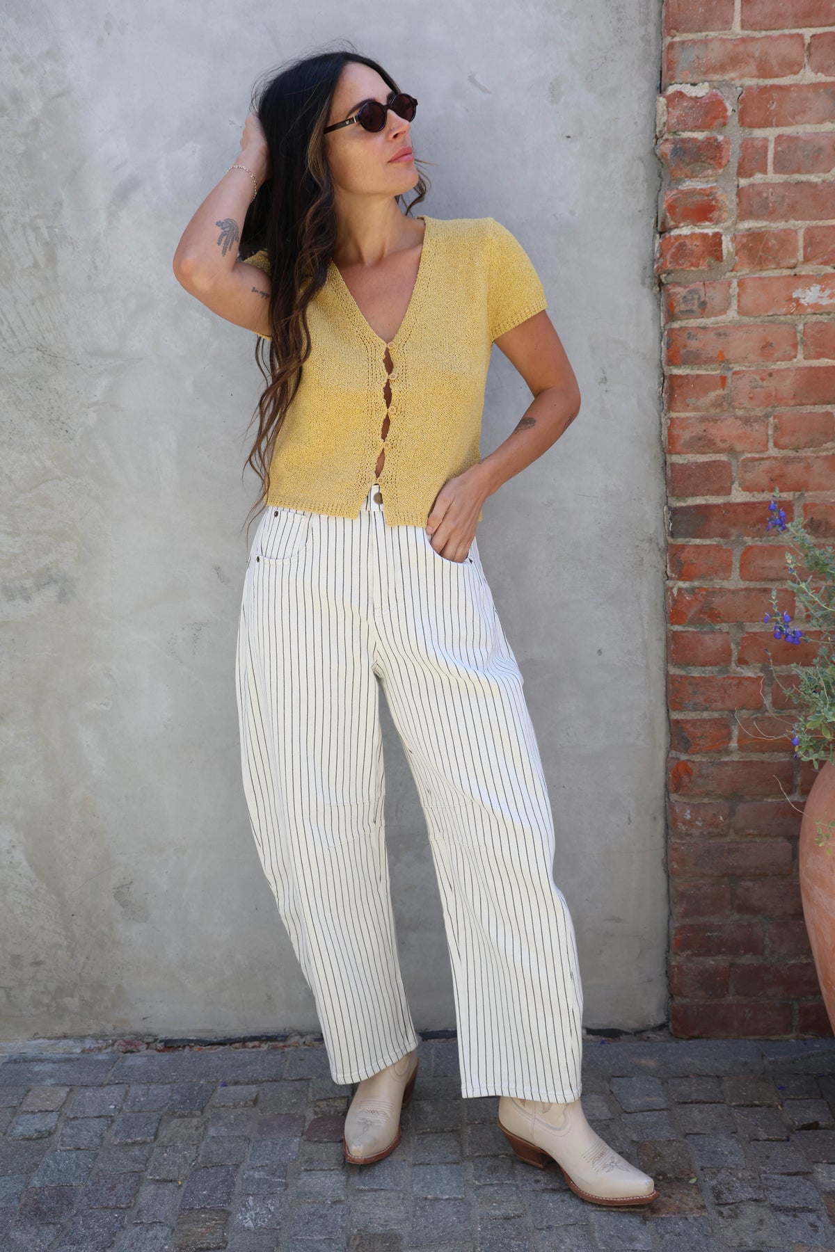 Woman wearing a yellow top and white striped pants standing against a brick wall.