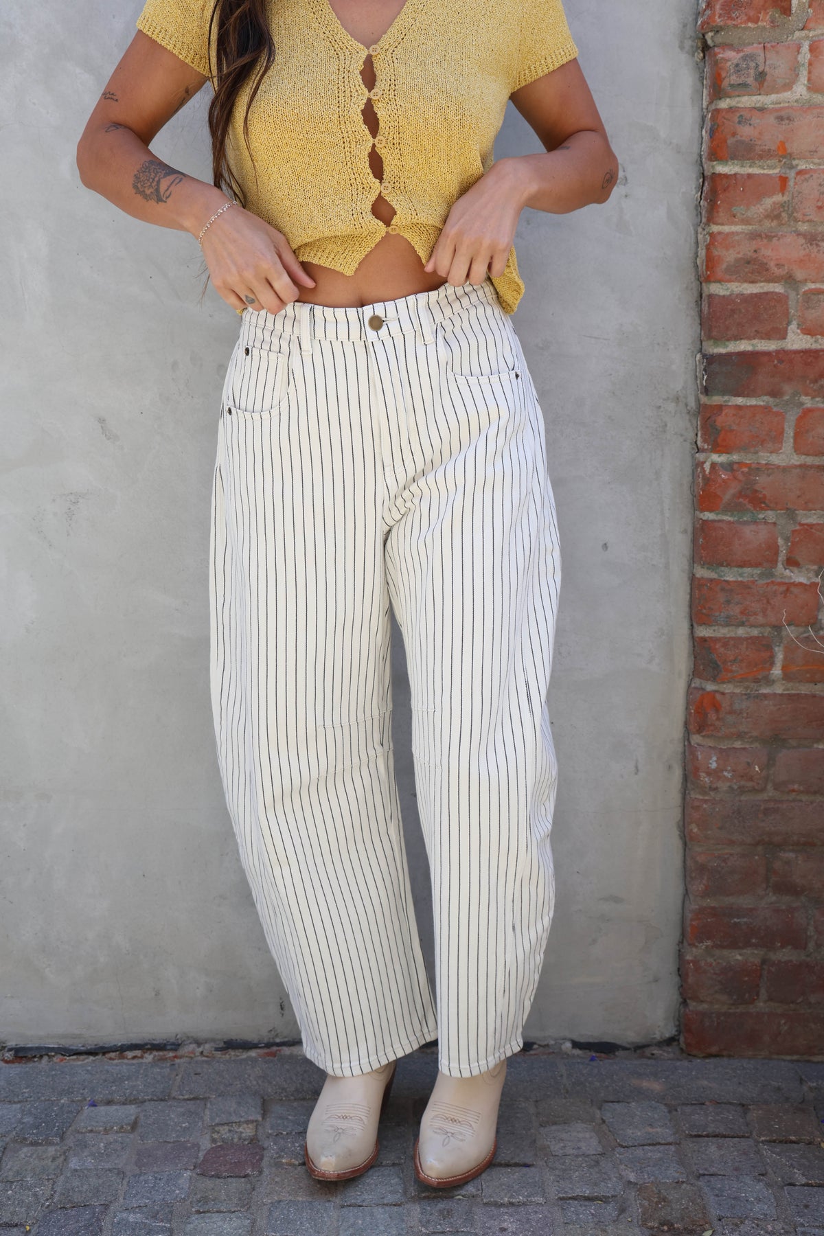 Person wearing a yellow knitted top and white striped pants standing against a brick wall.