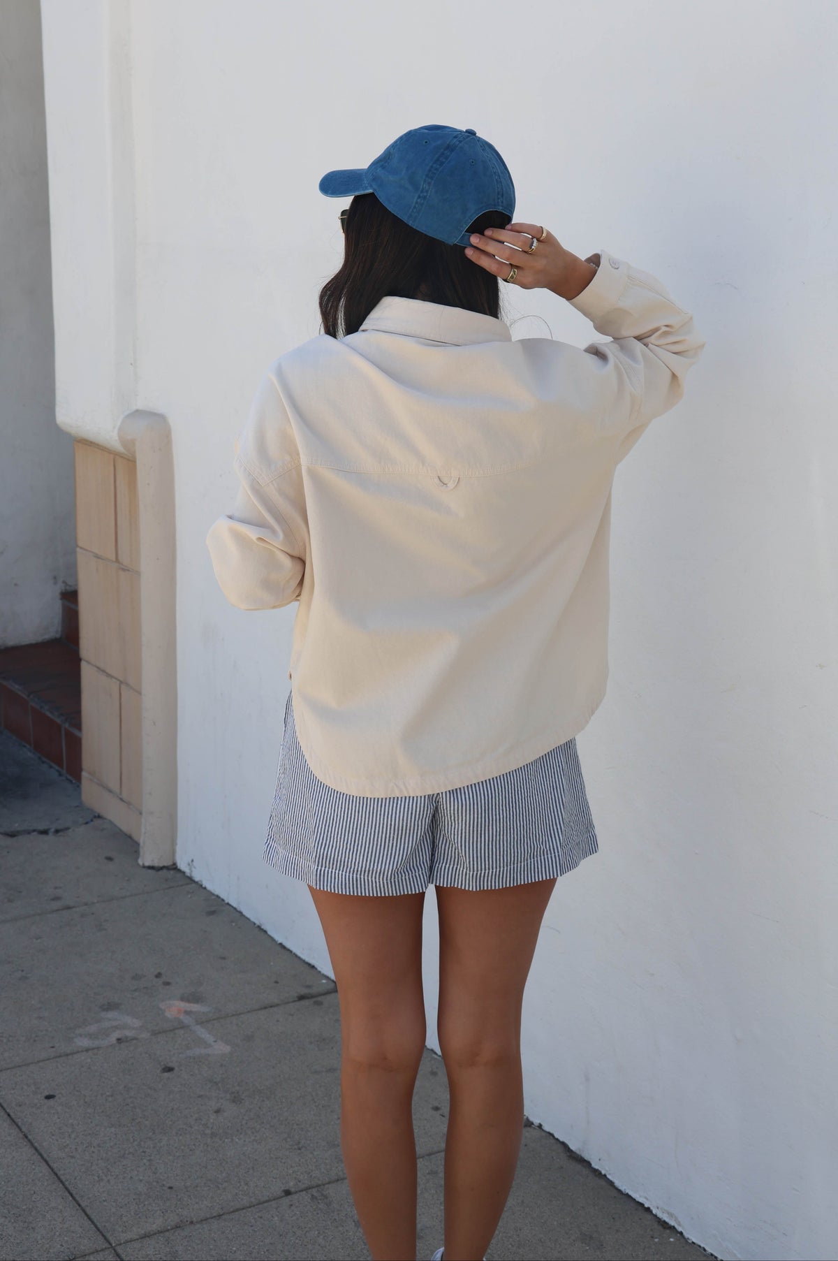 A woman seen from behind wearing a cream-colored button-up shirt, light gray shorts, and a blue baseball cap, standing against a white stucco wall.