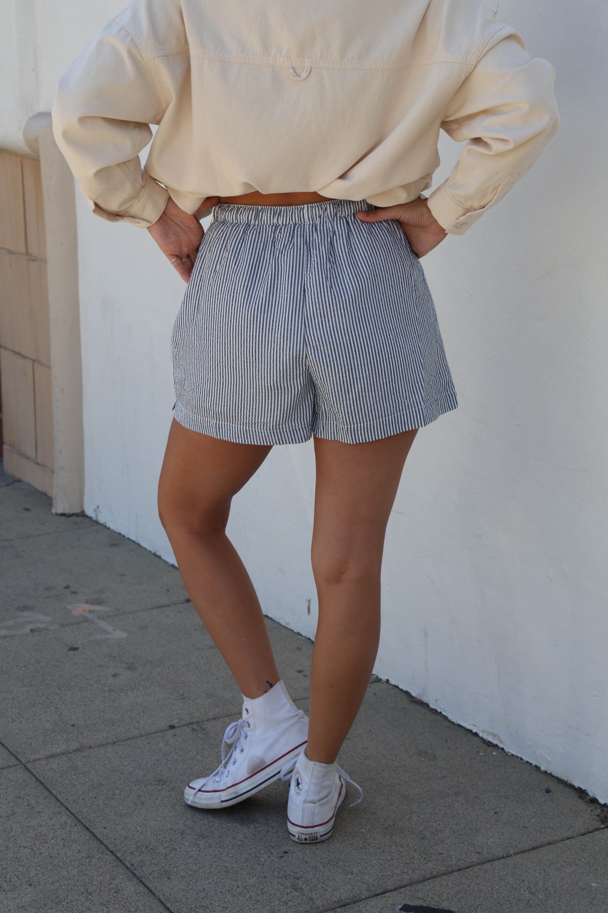 A person wearing light blue and white vertically striped shorts, a cream-colored long-sleeve top, and white sneakers, standing outdoors against a white wall.