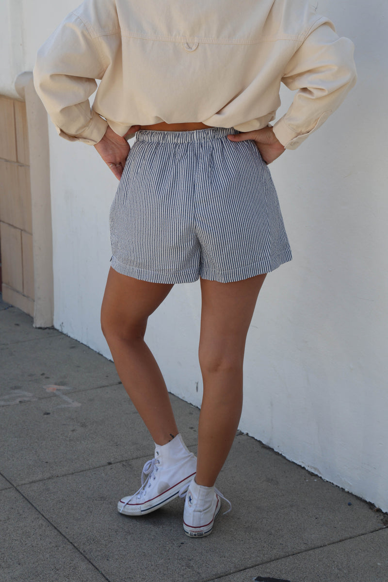 A person wearing light blue and white vertically striped shorts, a cream-colored long-sleeve top, and white sneakers, standing outdoors against a white wall.