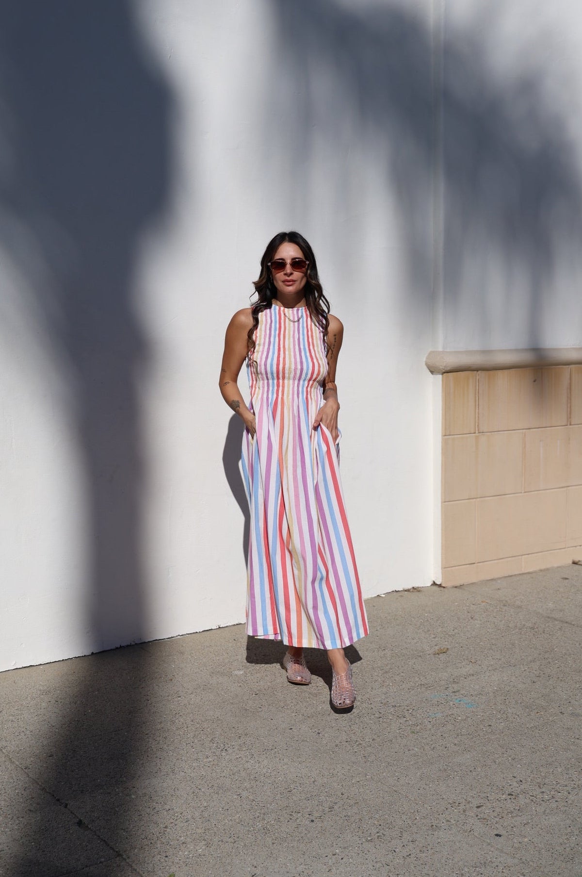 Woman in a colorful striped dress standing against a white wall with tree shadows.