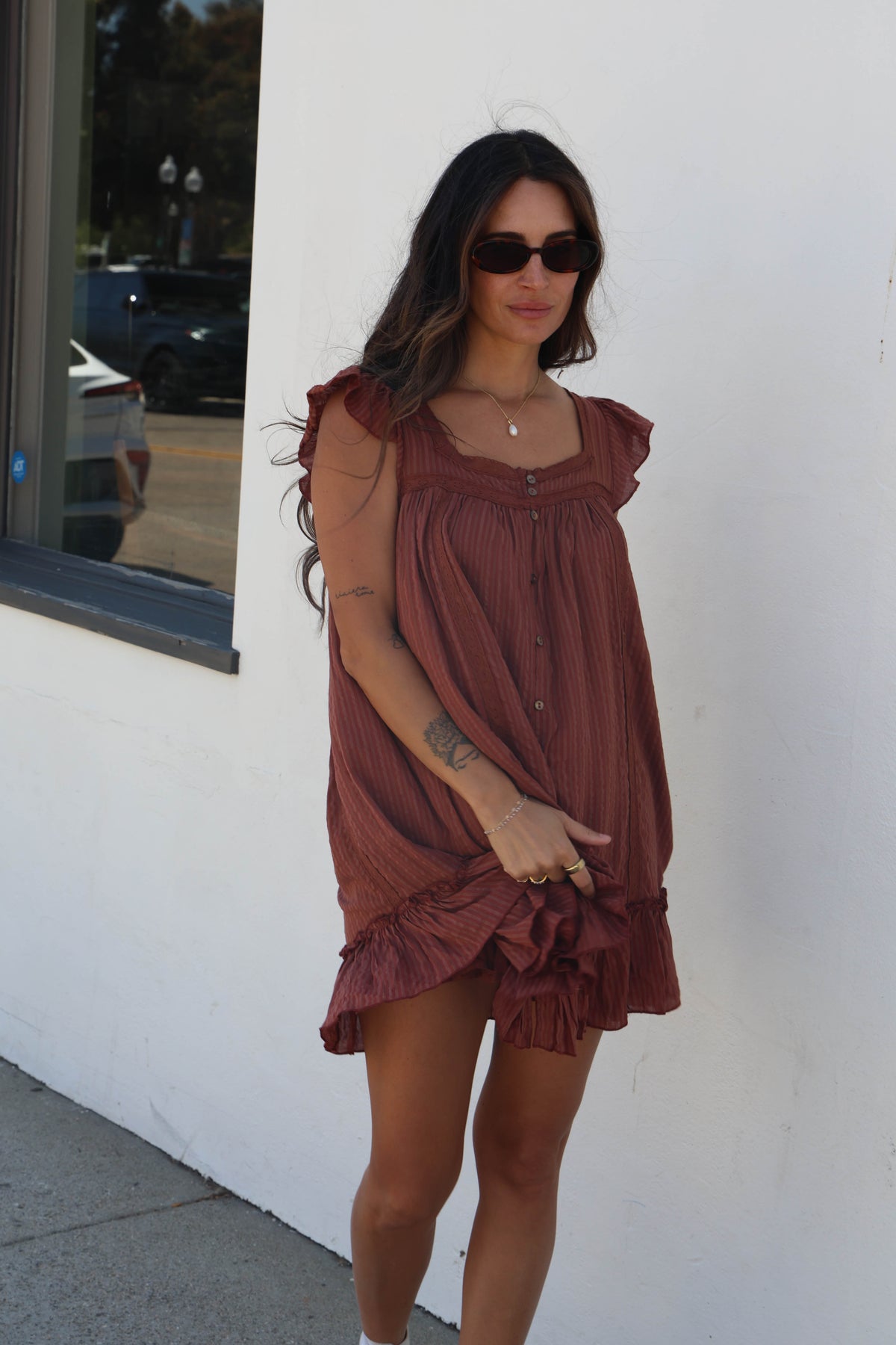 A woman wearing a rust-brown ruffled mini dress and black sunglasses, standing outdoors against a white wall.