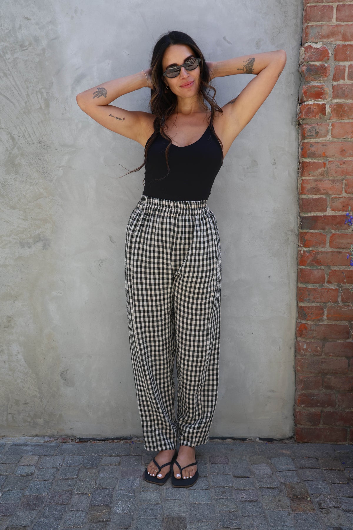 Woman wearing a black tank top and checkered pants against a textured wall.