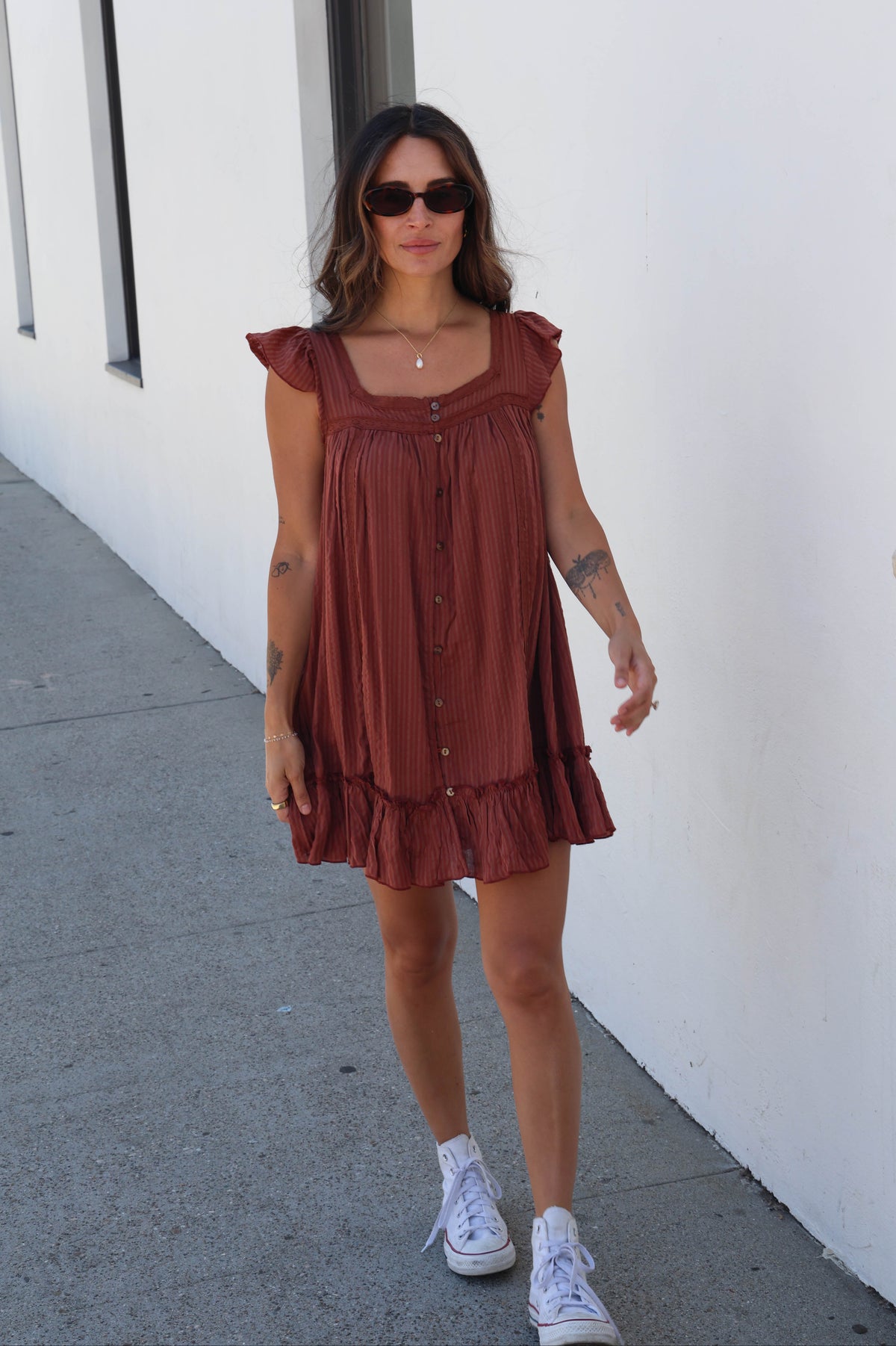 A woman wearing a rust-brown tiered mini dress with ruffle sleeves and front buttons, styled with white sneakers and sunglasses, walking along a sidewalk next to a white wall.