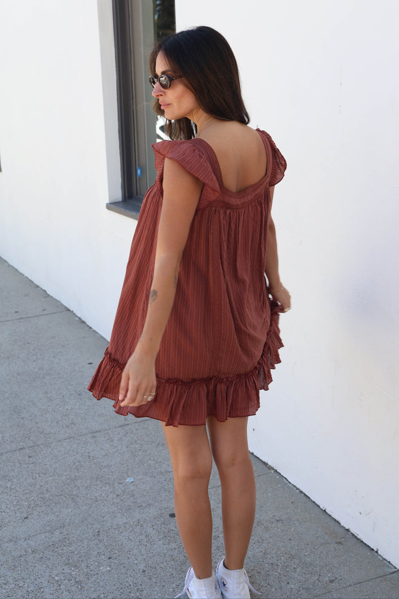 A woman wearing a rust-brown ruffled mini dress, standing outdoors against a white wall.