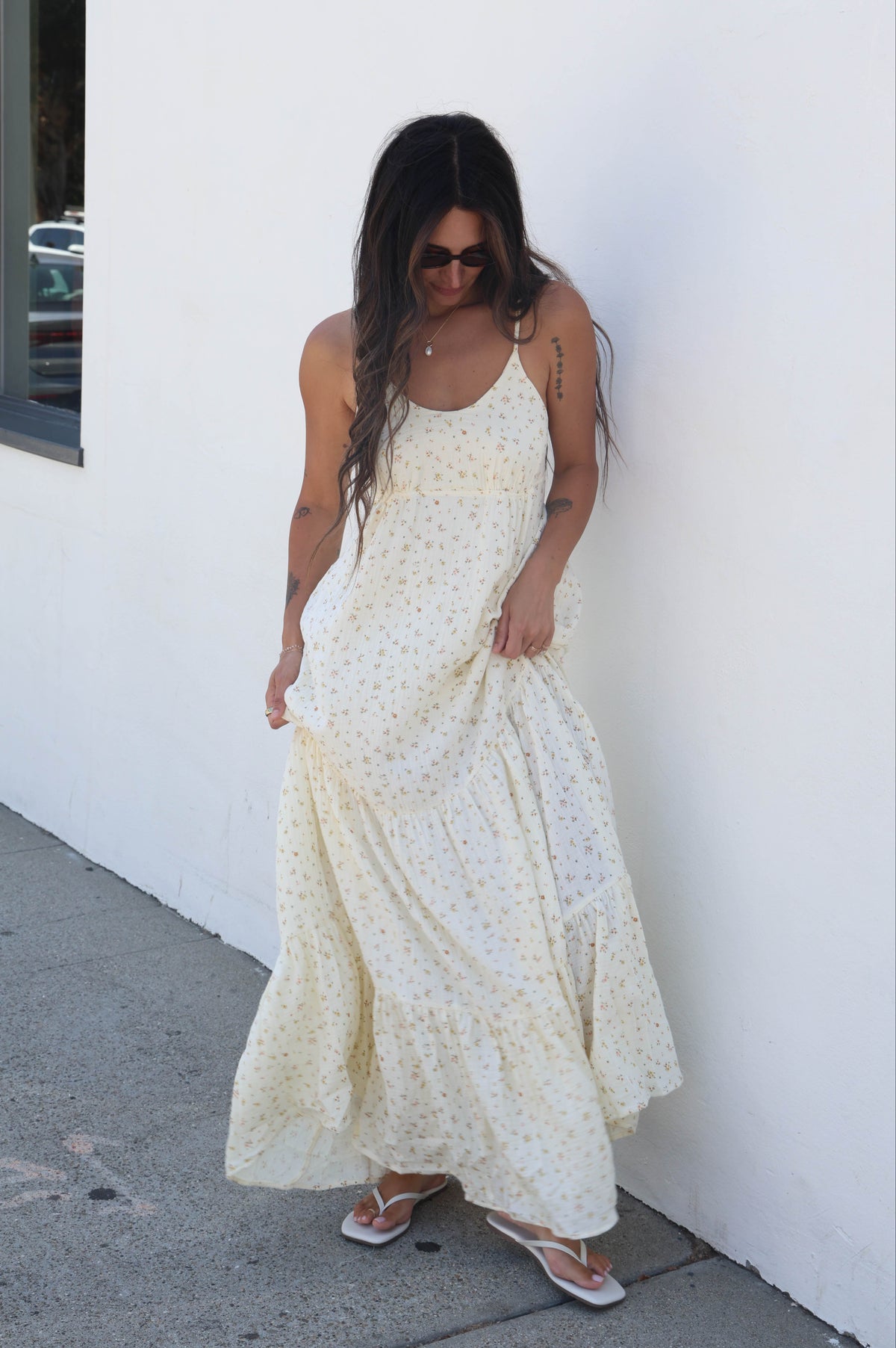 A woman wearing a cream-colored spaghetti strap maxi dress with a tiny yellow floral print, standing against a white wall.