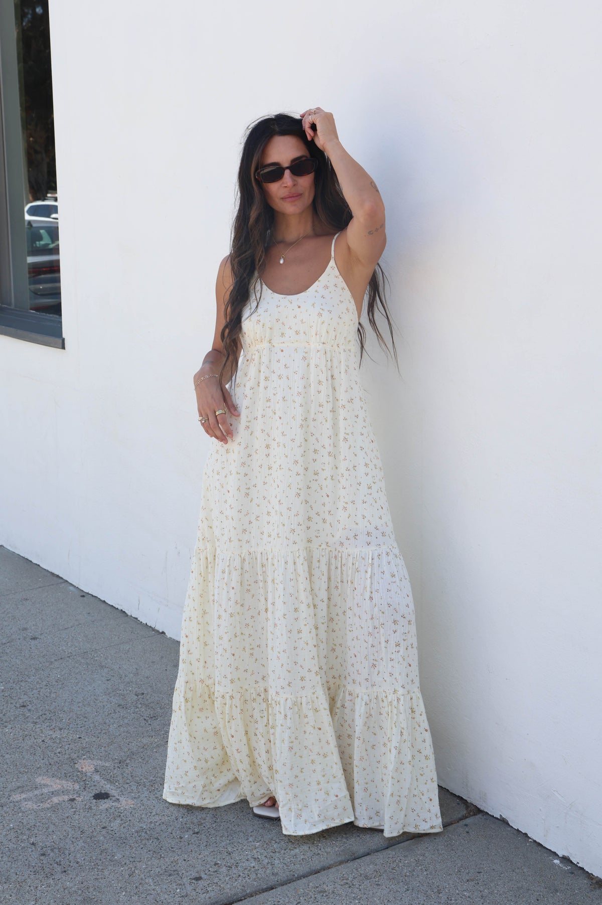 A woman wearing a cream-colored, tiered sleeveless maxi dress with a subtle white floral print, standing against a white stucco wall.