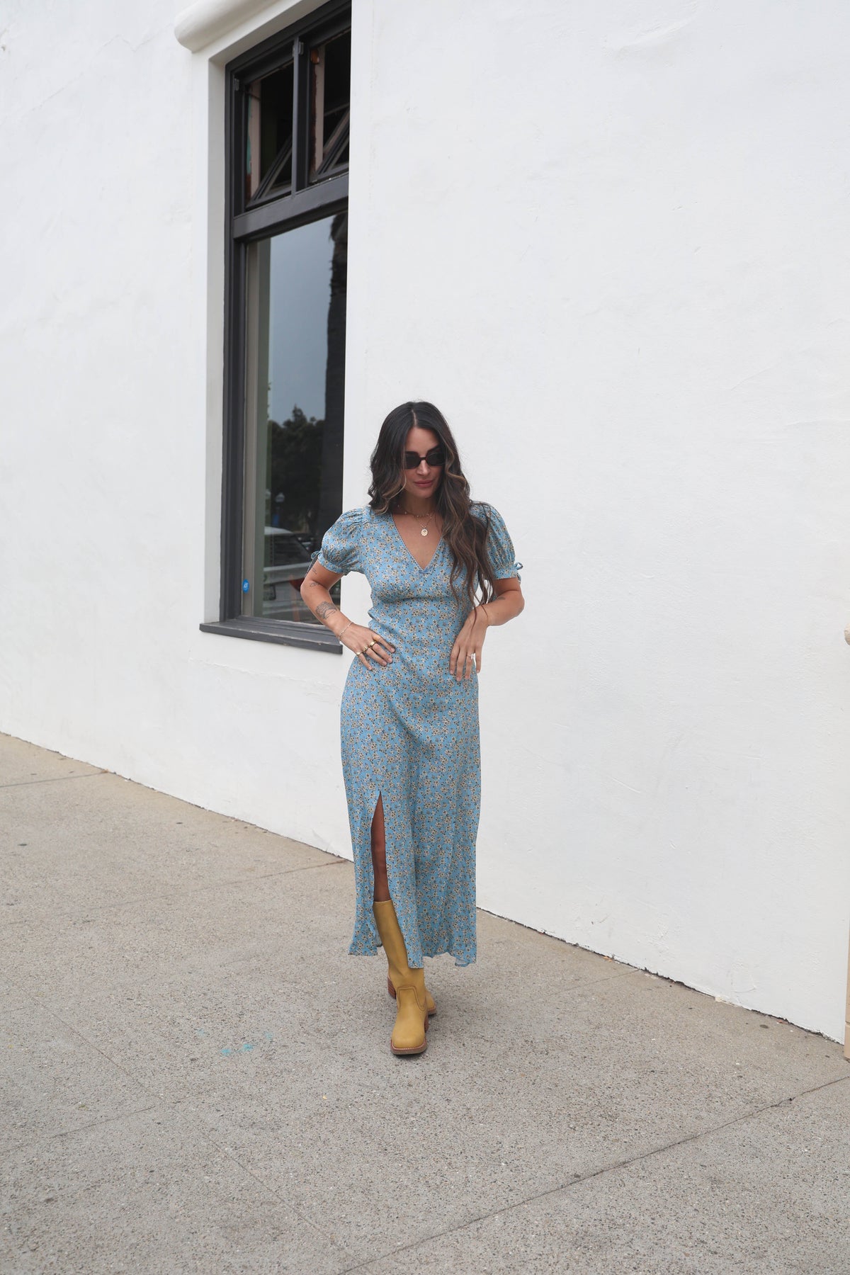 Woman in a light blue dress with yellow boots standing against a white wall.