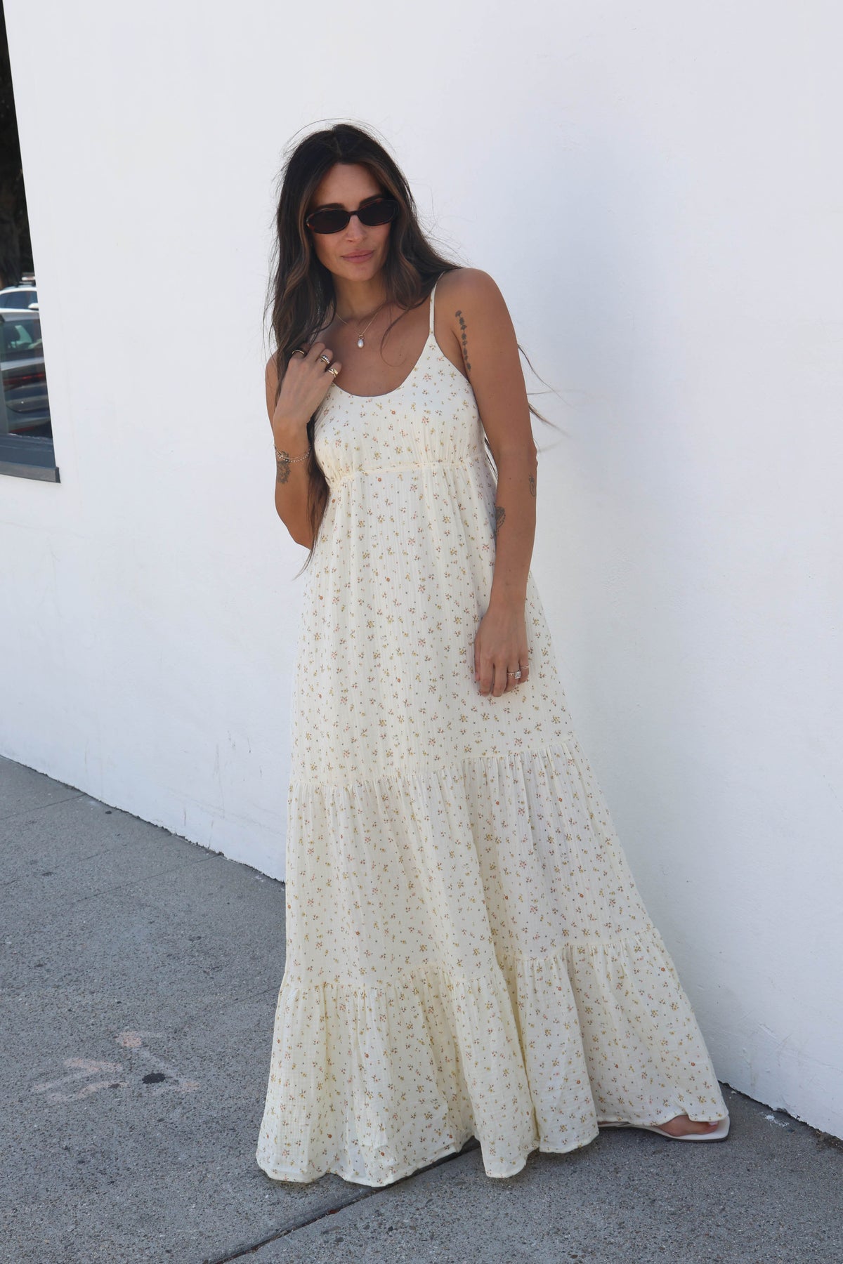 A woman wearing a cream-colored, tiered maxi dress with a small all-over floral print and sunglasses, standing against a white wall.