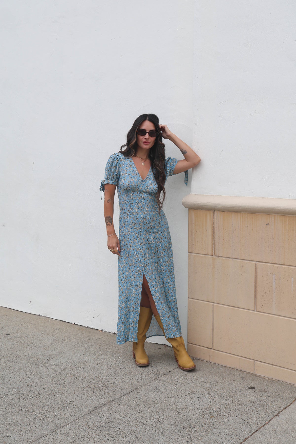 Woman in a blue floral dress and yellow boots standing against a white wall.