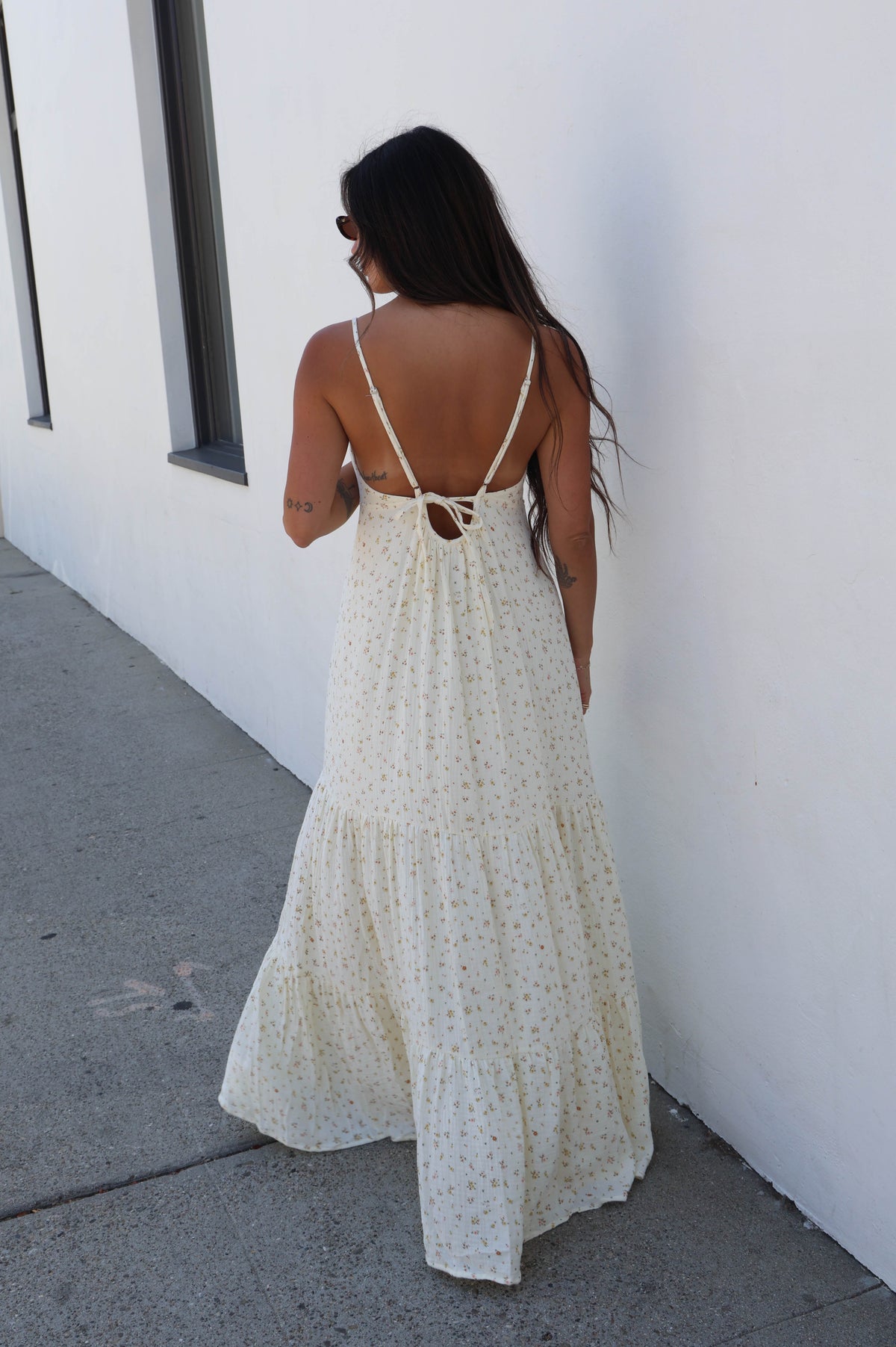 A woman wearing a cream-colored, tiered maxi dress with a small white dot pattern and an open back, photographed from behind as she walks along a sidewalk next to a white wall.