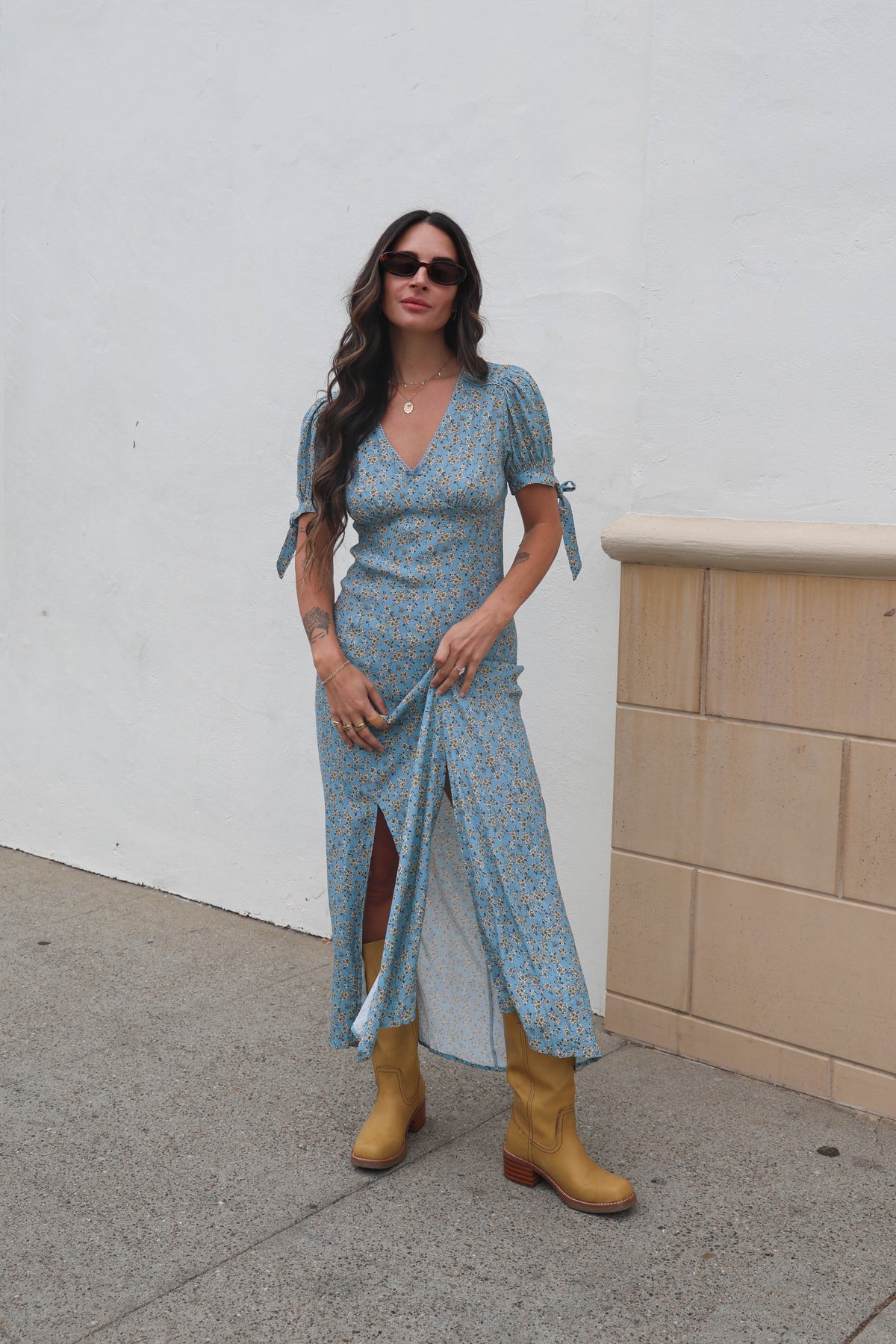 Woman wearing a blue patterned dress and yellow boots against a white wall.