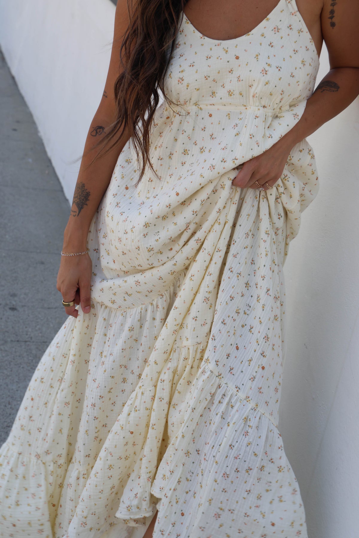 A woman wearing a cream-colored sleeveless maxi dress with a small all-over floral print, holding the skirt out to show its flow against a white wall.