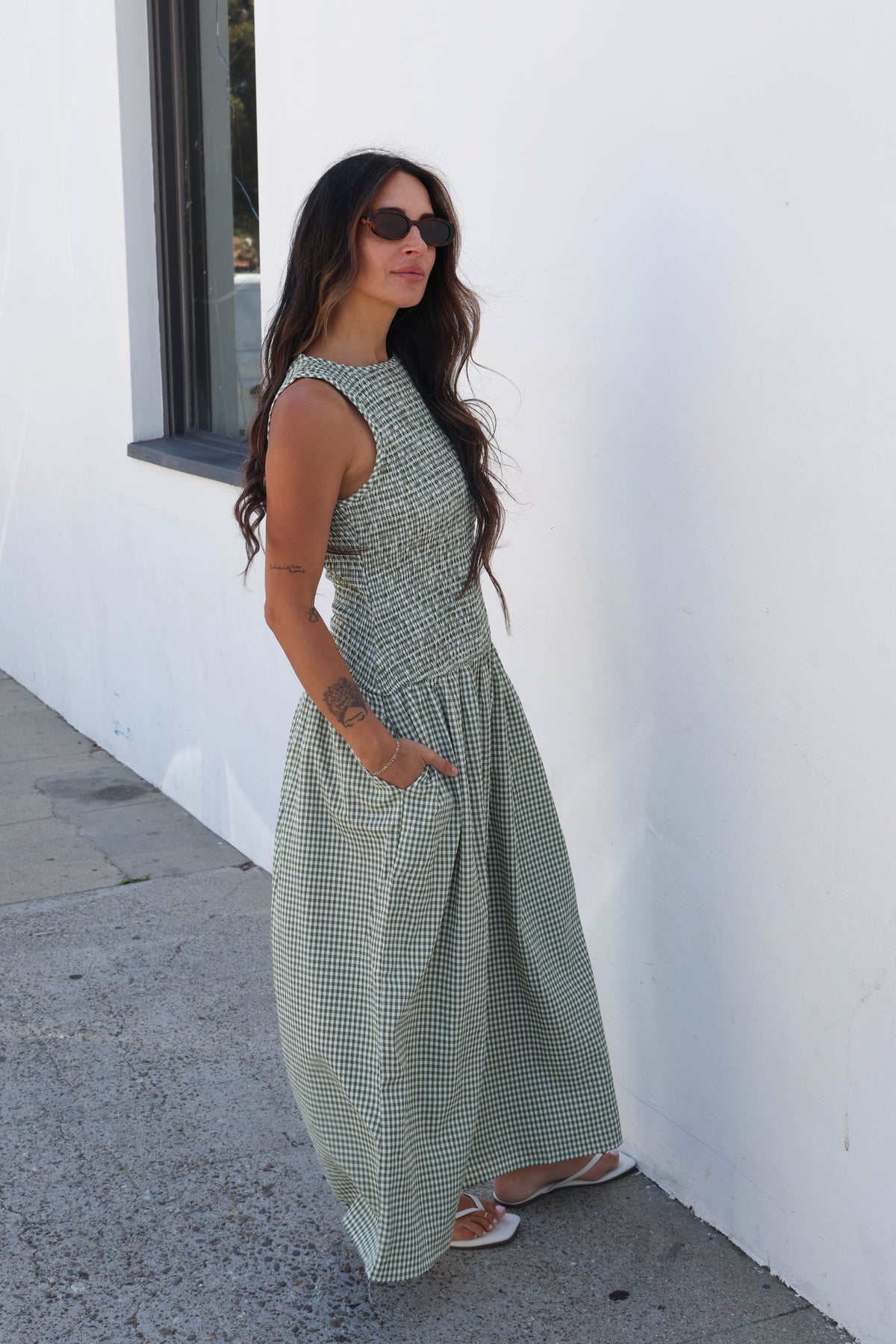 A woman wearing a sleeveless sage green and white gingham maxi dress with side pockets, standing outdoors against a white wall.