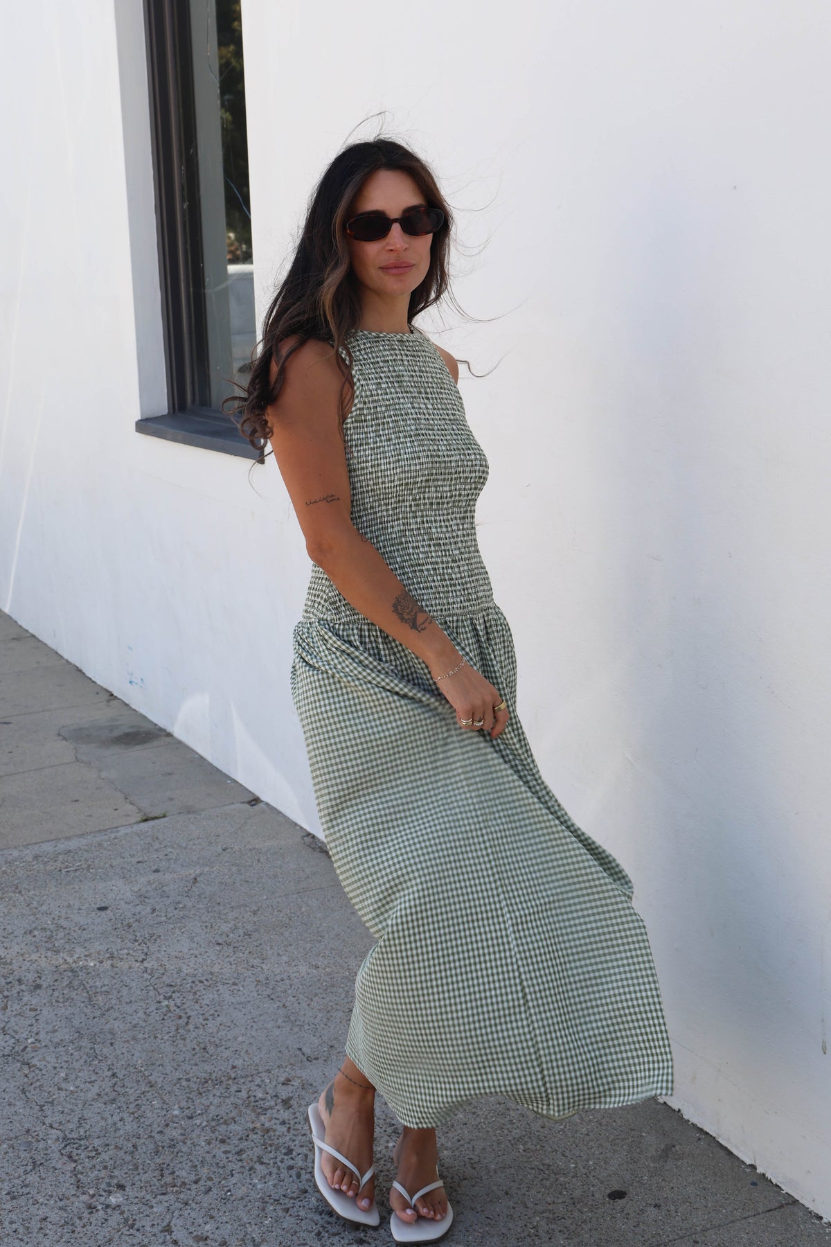 A woman wearing a sleeveless, high-neck, midi-length green and white gingham dress, styled with sunglasses and white sandals, standing outdoors against a white wall.