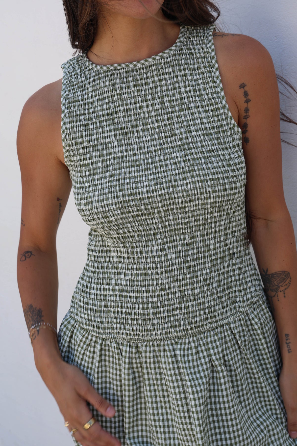 A close-up of a woman wearing a sleeveless smocked dress with a green and white gingham pattern, set against a plain white background.